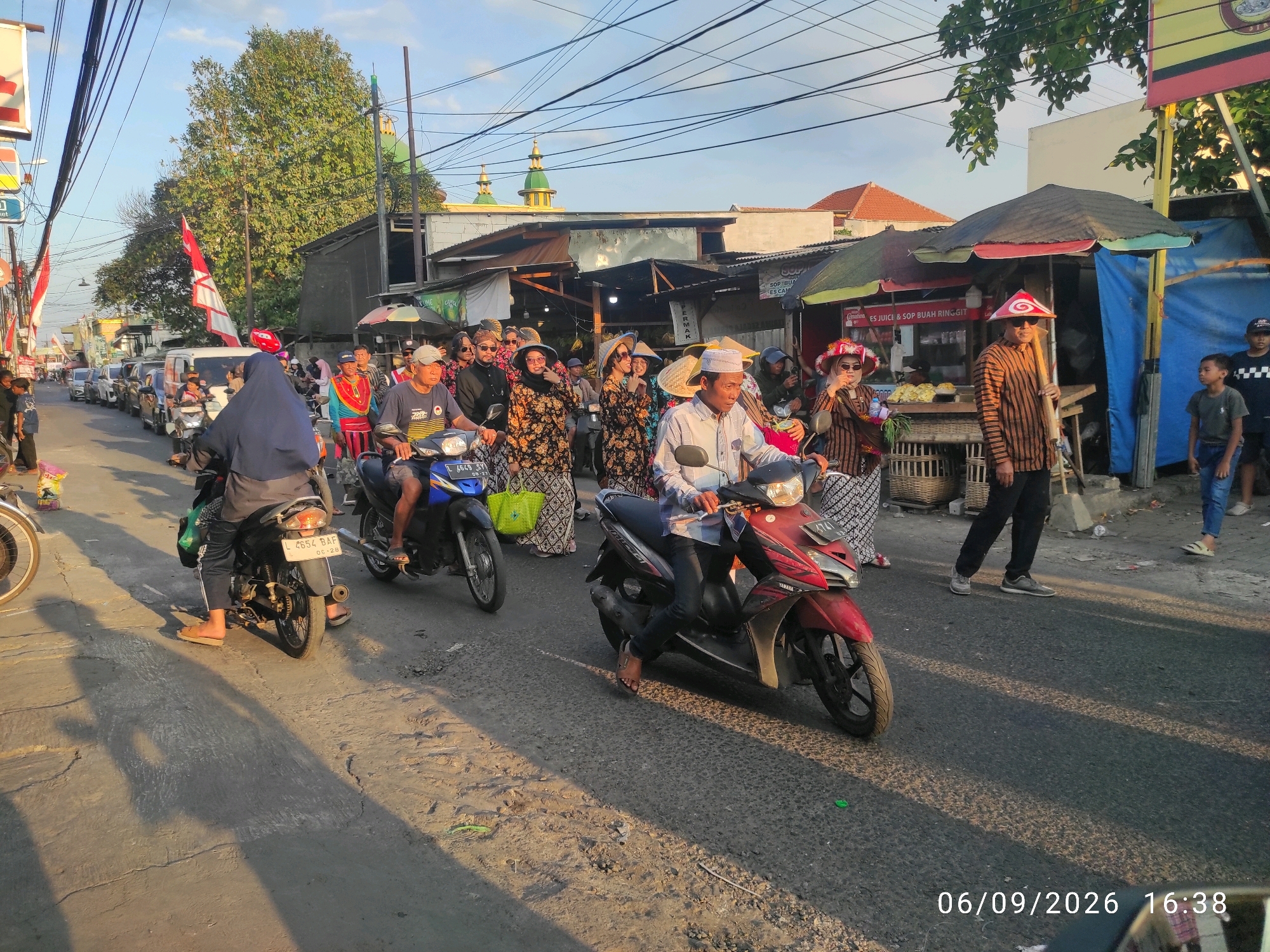This is a street-level, medium shot of a procession or parade in Wage, Indonesia. The street is paved and lined with shops and stalls. A line of motorcycles, some with riders and passengers, are moving down the street. People are dressed in traditional clothing, including batik patterns and conical hats. There are also some vendors and spectators present. The sky is clear and the sun is casting long shadows, indicating it is late afternoon. The overall scene depicts a lively cultural event.