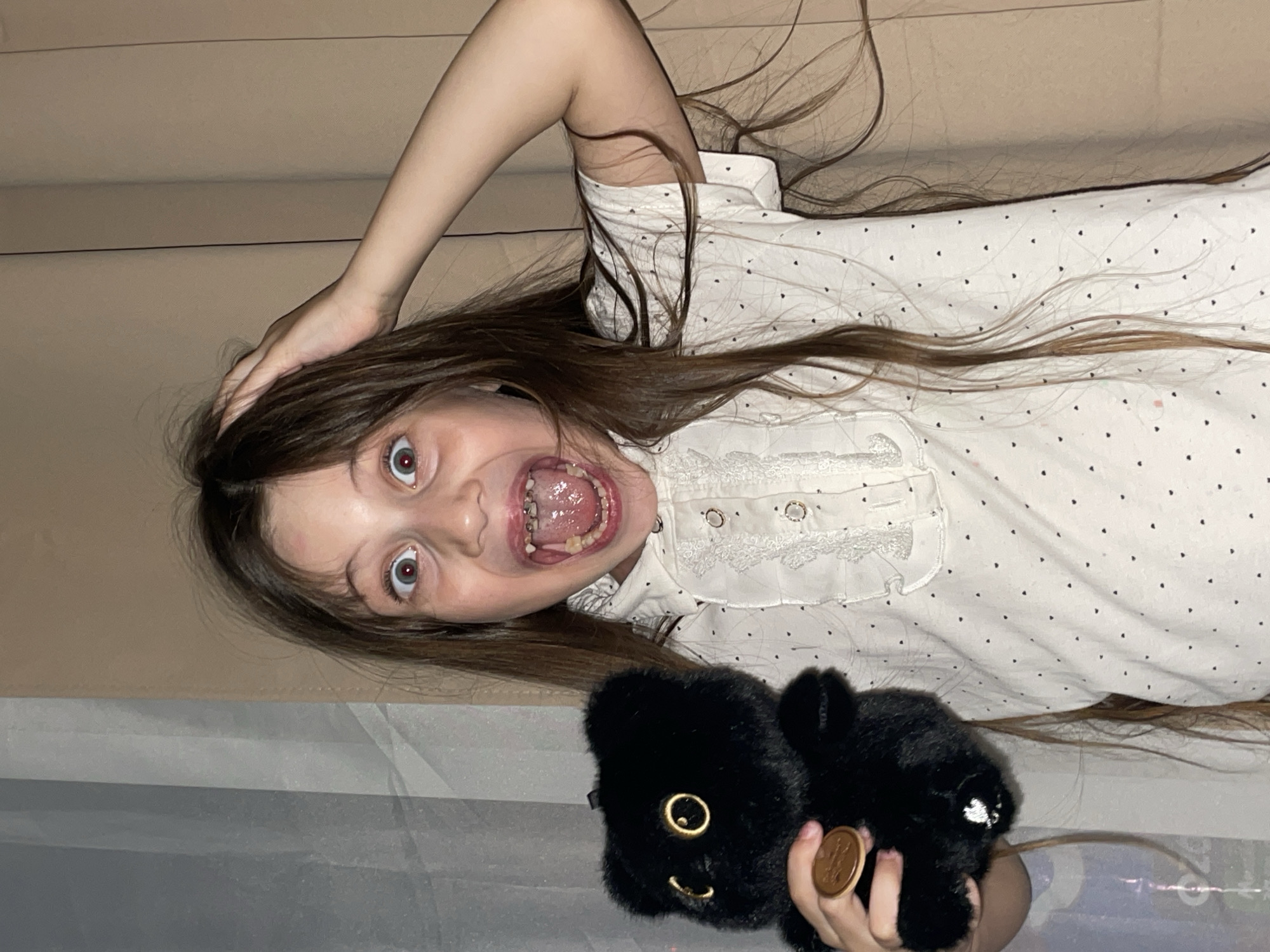 A young girl with long brown hair is lying on her back on a bed or soft surface. She is wearing a white shirt with small black dots. Her mouth is open wide, revealing her teeth, some of which appear to have dental work. Her eyes are wide and expressive. She is holding a small, black stuffed animal with golden eyes and a smiling mouth. The environment appears to be indoors, possibly a bedroom, as evidenced by the bed and soft lighting. There is no indication of Izhevsk, Russia or other location information visible in the image.