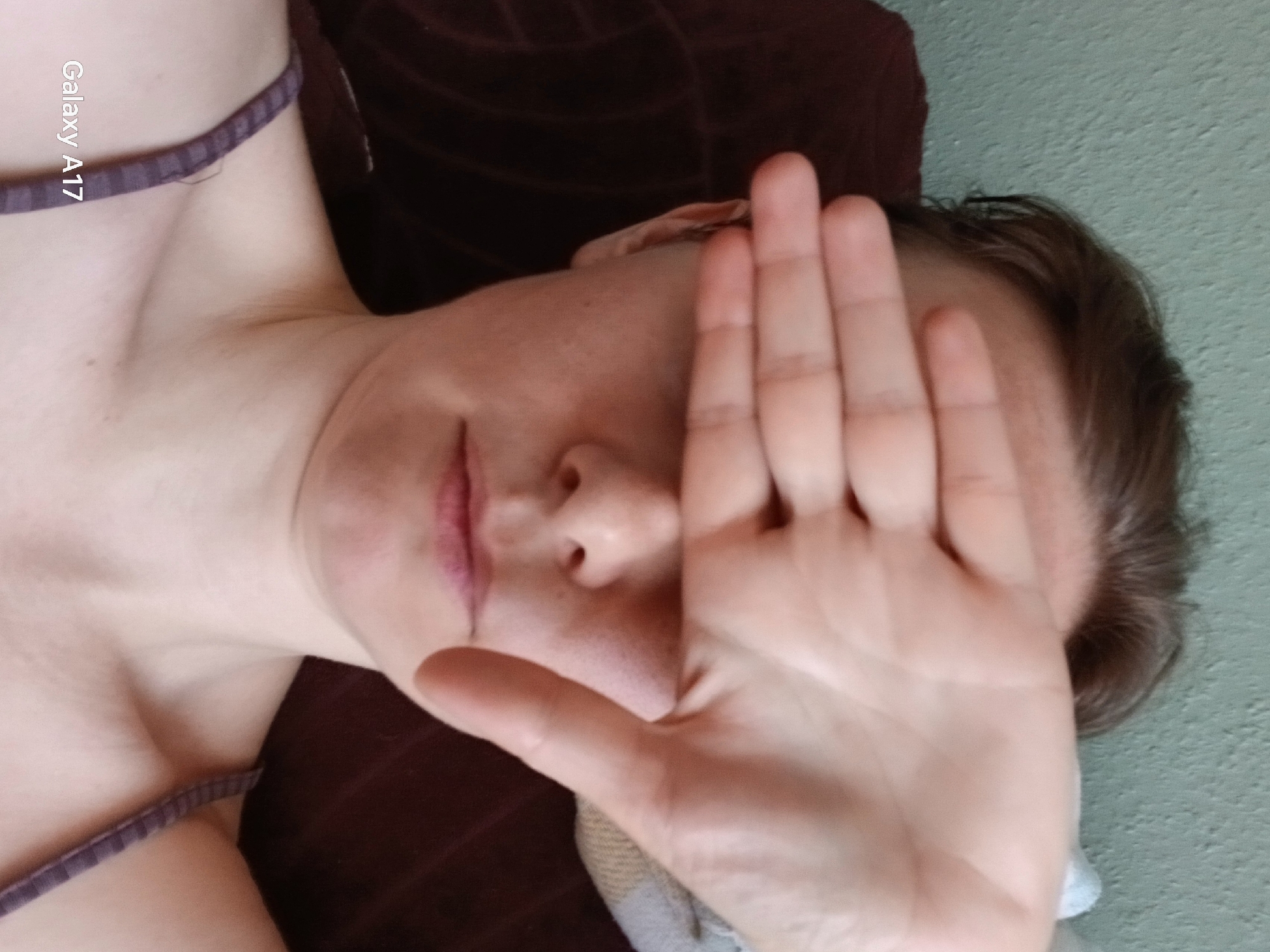 A fair-skinned person with short brown hair is lying down with their right hand covering their face. The person is wearing a thin-strapped top with a dark purple band. The person is lying on a dark brown, possibly velvet-textured, surface. A wall or surface with a light green and textured appearance is visible to the right of the person's head. The inscription "Galaxy A17" is visible in the upper left of the image. There is no indication of the location being Yekaterinburg, Russia.