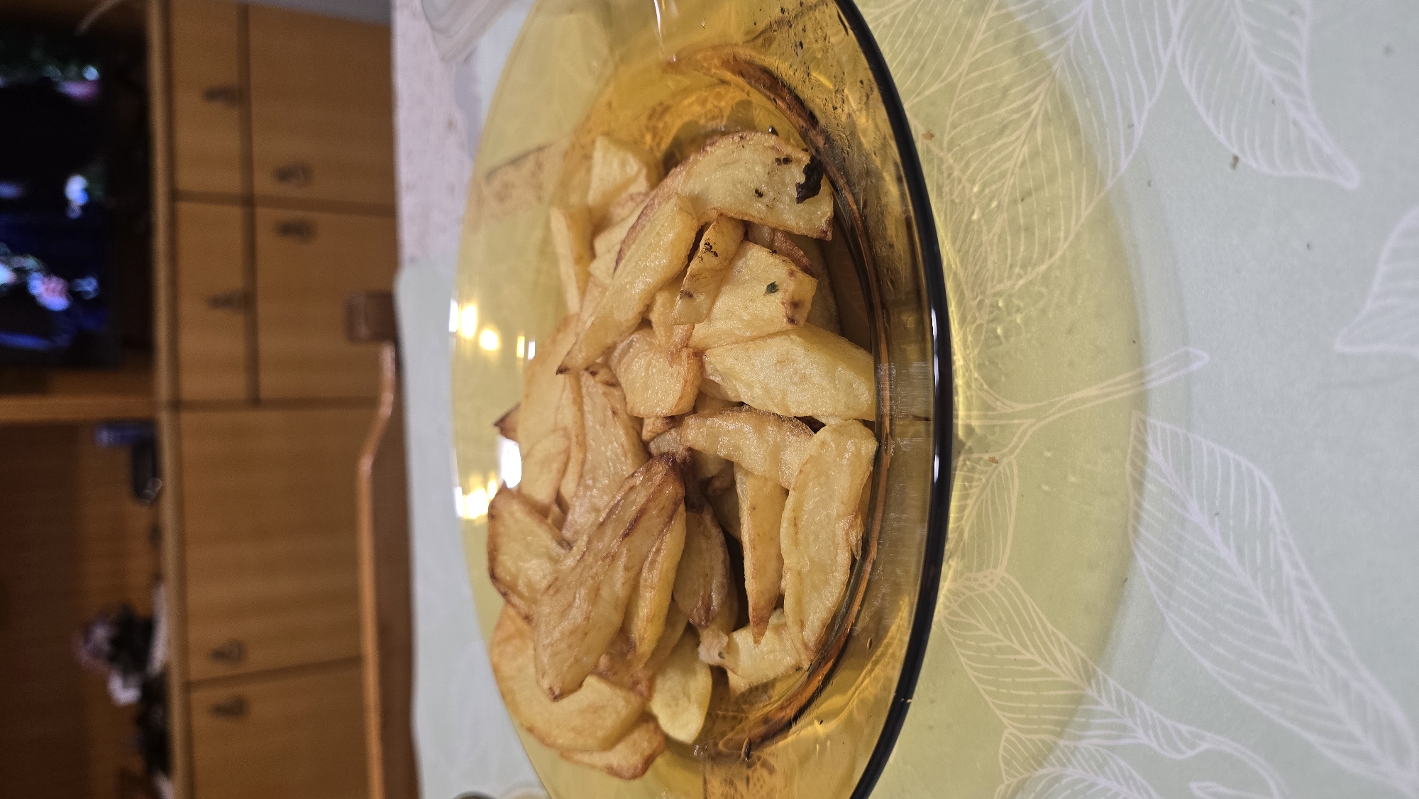 A close-up shot shows a glass plate filled with golden-brown french fries. The plate is made of amber-colored glass and sits on a light green tablecloth with a white leaf pattern. In the background, a wooden cabinet with multiple drawers is visible, and a television screen displays an image. The scene appears to be indoors, likely in a residential setting in the city of Troyan, Bulgaria, as suggested by the context. The lighting is dim, with some reflections visible on the plate and in the background.