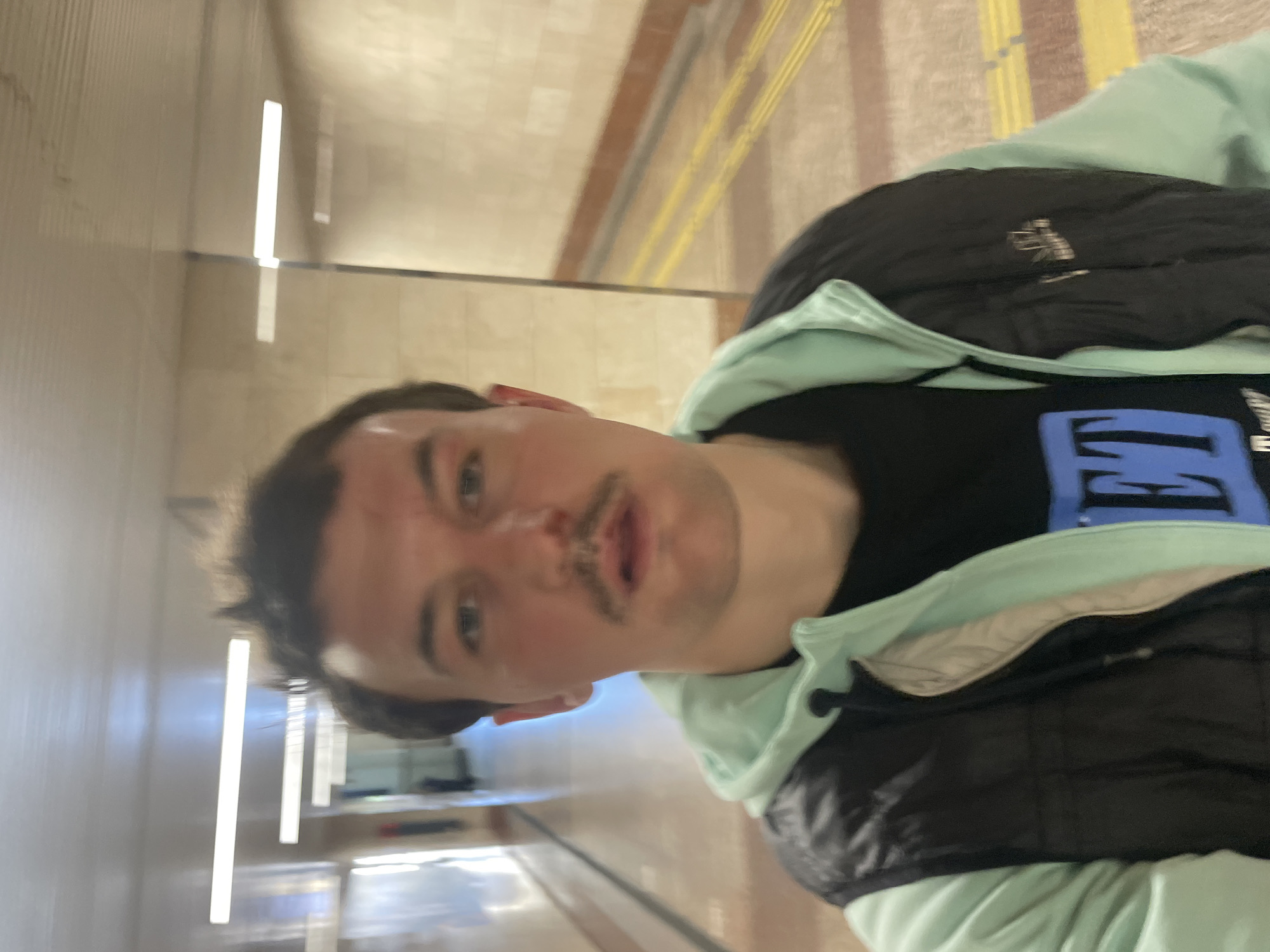 A person is in what appears to be an indoor hallway or tunnel setting. The walls are light in color, and there are linear light fixtures on the ceiling. Yellow and brown stripes are visible in the background. The person has short dark hair and a mustache and is wearing a dark shirt with a light blue graphic, a light green hoodie, and a black puffer vest. The location appears to be an underground passage based on the lighting and architectural design. Without more context, determining the exact location in Sochi, Russia, is not possible.