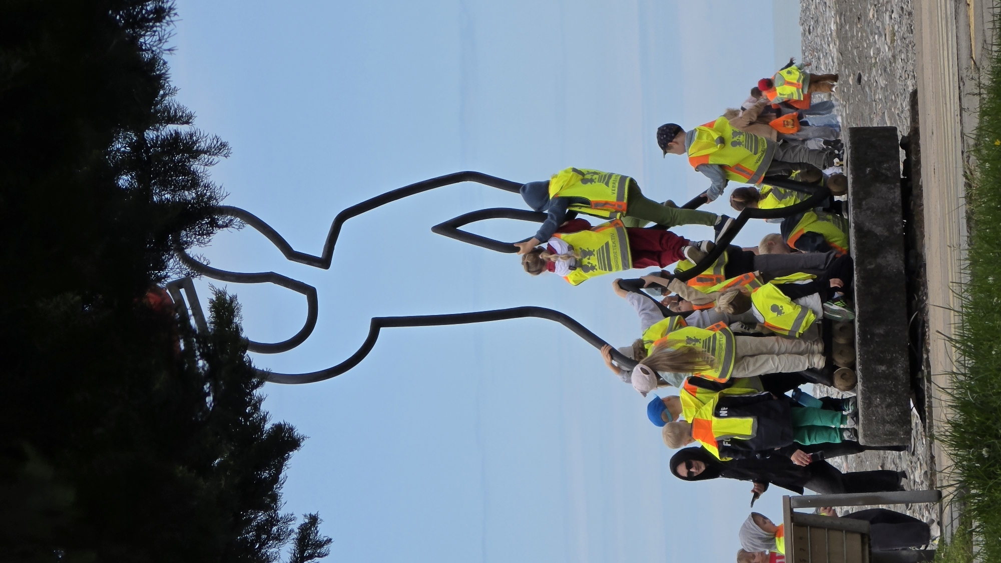 The image depicts a group of children in bright yellow safety vests interacting with a large, abstract metal sculpture in Batumi, Georgia. The sculpture, situated near a tall evergreen tree, appears to be a stylized representation of a human figure with outstretched arms. The children, mostly wearing casual clothing, are positioned along the sculpture, some holding onto the metal arms, suggesting play or interaction. The backdrop is a clear blue sky. The scene appears to be outdoors near a pedestrian walkway with a gray concrete barrier and a patch of grass.