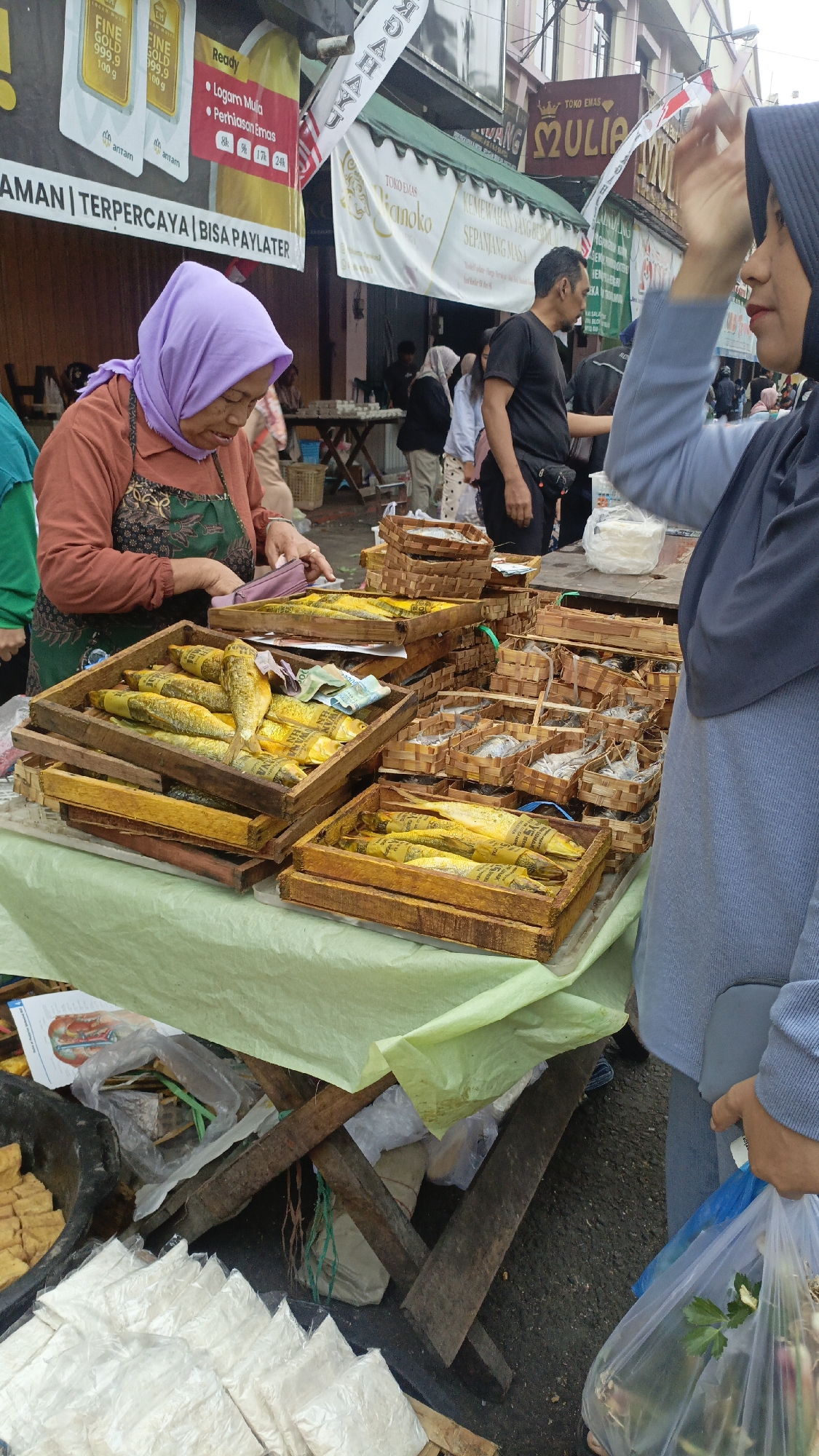 In a bustling outdoor market in Salatiga, Indonesia, a vendor wearing a lavender headscarf and an apron is arranging fish on a table covered with a pale green cloth. The fish, some wrapped in yellow paper, are displayed in wooden crates. Behind the vendor, other people are visible, including a man in a black t-shirt and a woman in a blue hijab holding a plastic bag filled with produce. The market appears to be situated on a street with buildings in the background displaying various signs, including one for "TOKO EMAS MULIA," indicating a gold shop. The ground is paved, and the overall scene suggests a typical Indonesian street market environment.