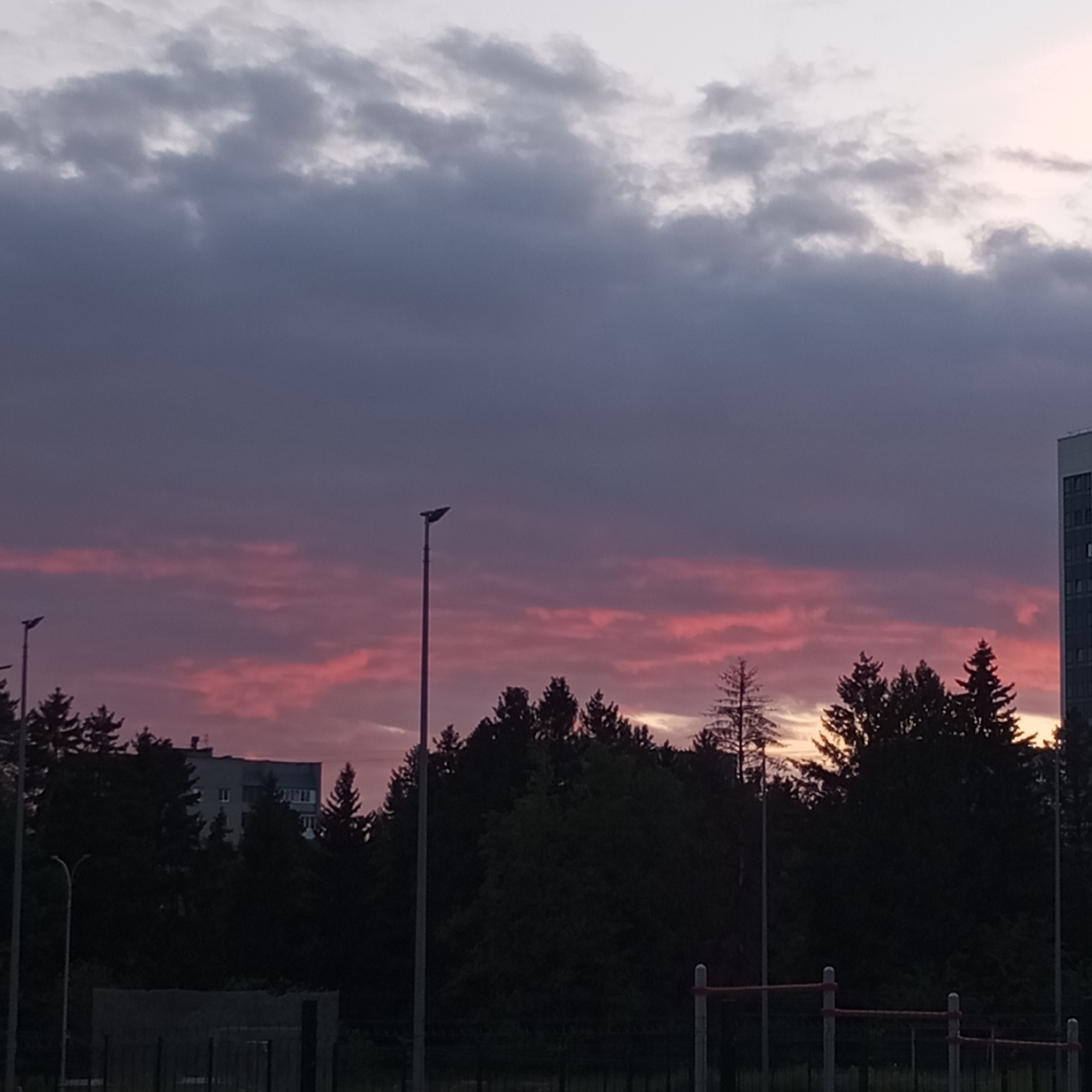 The image captures a dusky sky over a city with a hint of pink and purple hues, indicating either sunset or sunrise. Silhouetted against this sky are a dense line of trees, a few streetlights, and the upper floors of a tall building on the right. In the foreground, there are elements of an outdoor recreational area, possibly a sports ground, with horizontal bars supported by vertical poles. The scene appears to be a typical urban landscape, perhaps a park or a residential area, with the sky dominating the upper portion of the frame. The presence of apartment buildings and streetlights suggests an urban environment. While the specific location within Izhevsk, Russia, cannot be pinpointed without additional information, the overall aesthetic points to a developed city. There are no people visible in the image.
