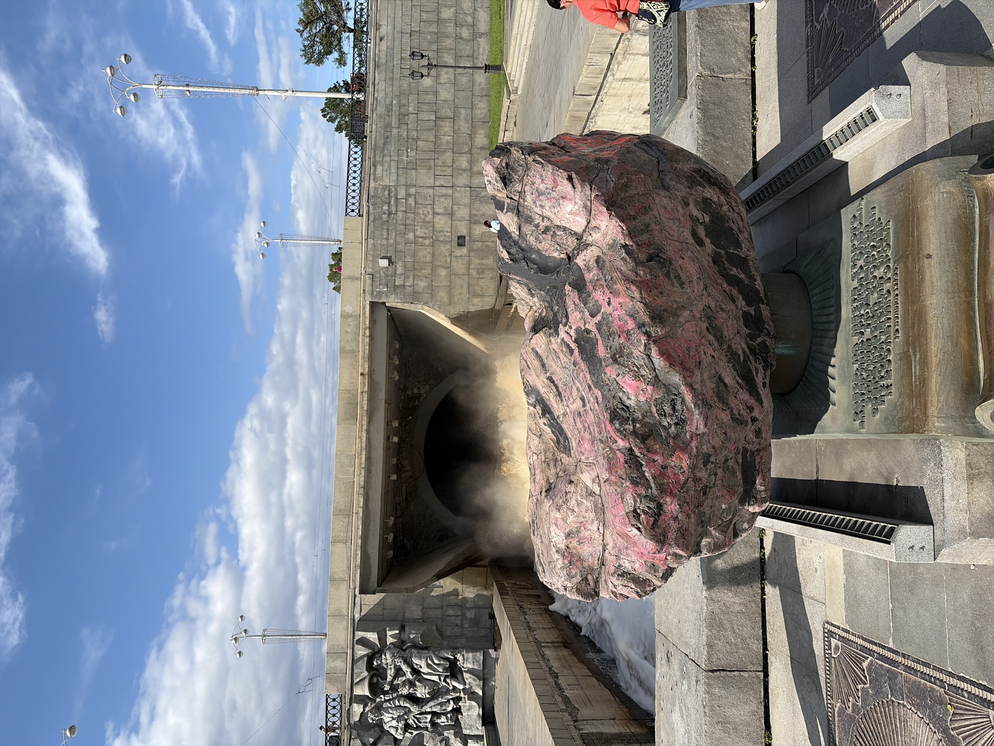 The image depicts the "Kazantsev's Stone" sculpture, also known as the "Stone Flower," located at the City Pond embankment in Yekaterinburg, Russia. The sculpture is a large, roughly spherical rock painted with red and black patterns, resting on a bronze pedestal with inscribed text. In the background, water is forcefully exiting an arched tunnel beneath a concrete bridge, creating a frothy spray. To the left of the tunnel is a stone relief depicting several figures. The environment is outdoors under a partly cloudy blue sky, with lampposts and trees visible in the distance. A few people are present in the scene, including one woman in an orange shirt in the foreground and another person walking near the bridge in the mid-ground. The overall scene suggests a public outdoor space, likely a park or promenade, with a focus on the prominent sculptural artwork and the dramatic water feature.