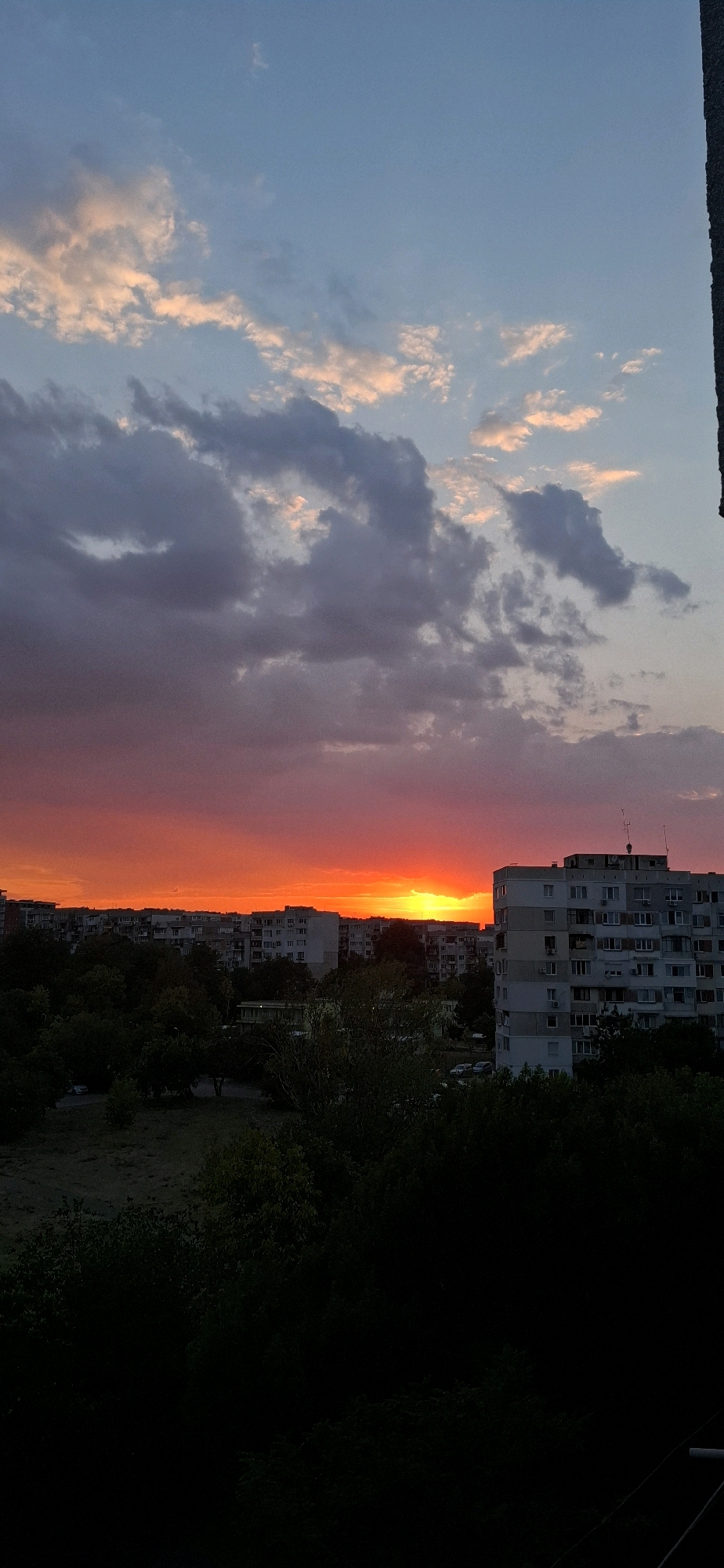 This is a vertical, wide-angle photograph taken from a high vantage point, likely a balcony or rooftop, overlooking a city landscape at sunset. The sky dominates the upper half of the frame, transitioning from a pale blue at the top to a vibrant orange and red glow near the horizon, indicative of a sunset. Scattered clouds, tinged with pink and gray, are visible in the sky, adding texture and depth.
Below the sky, a dense line of apartment buildings stretches across the mid-ground. These buildings are multi-story, with a consistent architectural style suggesting a Soviet-era urban development, common in many Eastern European cities. Some buildings have visible windows and balconies. To the right, a larger, modern apartment building stands out with its grey facade and multiple floors.
In front of the apartment buildings and extending to the lower part of the image is a dark expanse of trees and foliage, obscuring the ground level. The trees are lush and dense, casting the foreground into deep shadow. A few cars are sparsely visible parked on a street between the trees and buildings, suggesting a residential area. The overall atmosphere is tranquil, captured during the golden hour.
The image was taken in the Mladost district of Sofia, Bulgaria.