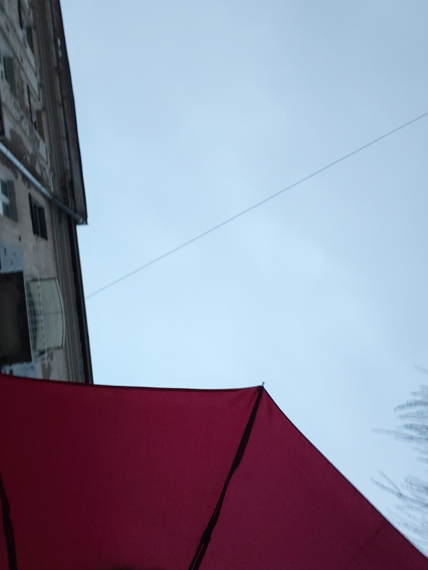A low-angle, outdoor shot shows the corner of an old building, a red awning, an overhead wire, and the sky. The building, located on Pushkinskaya Street in Izhevsk, Russia, is multi-storied, weathered, and has several visible windows, one of which has a wire cage. A single, horizontal wire crosses the upper part of the frame against a backdrop of pale blue sky. The foreground is dominated by a triangular red awning. In the lower right corner, there is a suggestion of bare tree branches, slightly out of focus.