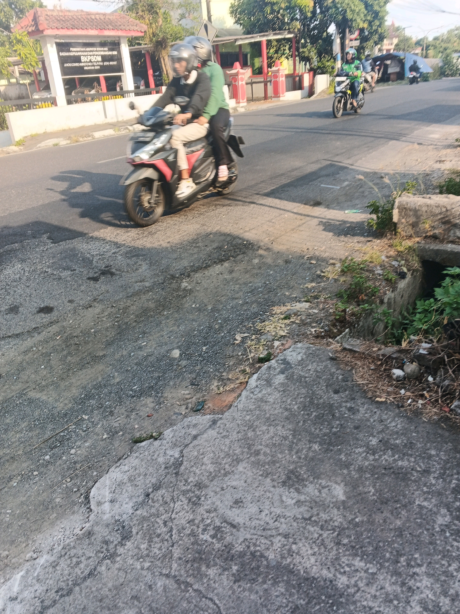 In Ungaran, Indonesia, two individuals wearing helmets ride a red and black motorcycle on a paved road. The rider in the front wears a black jacket and light-colored pants, while the passenger behind wears a green jacket and dark pants. To the left of the road, a building with a red roof and white walls is visible, bearing a sign that reads "PEMERINTAH KABUPATEN SEMARANG" and "BKPSDM". Behind the building, trees and other structures are present. Other motorcycles are seen in the background on the road. The foreground of the image shows a rough, gray surface, possibly concrete or asphalt, with some scattered debris and vegetation. The lighting suggests it is daytime, with shadows cast on the road.
