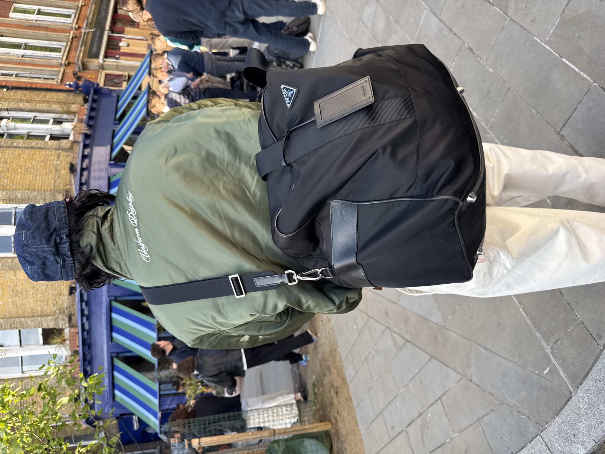 The image captures a casual street scene in an urban environment, consistent with London, United Kingdom, seen from a slightly elevated perspective looking down. The central subject, viewed from behind, is a person wearing a sage green bomber jacket with white cursive text on the back, which appears to read "Uniform Drip." They also wear light-colored, possibly white or cream, trousers and a dark blue denim hat or beanie.
A prominent black Prada duffel bag or holdall is slung over the person's right shoulder. The bag features the recognizable Prada triangular logo, with "PRADA" clearly visible in white text, and an attached rectangular leather tag also embossed with "PRADA." The bag has black straps and silver-toned hardware.
In the blurred background, other individuals are visible, suggesting general street activity. Behind them, classic brick buildings with multiple windows line the street. A distinctive blue structure with diagonal blue and light blue stripes, possibly an awning or part of a market stall, adds a vibrant element to the background. Some green foliage is also present on the left side of the frame.
The ground is paved with grey flagstones, typical of a city sidewalk or square. The lighting indicates it is daytime, likely under clear skies, with natural light illuminating the scene. The overall atmosphere is that of a bustling, everyday urban moment.