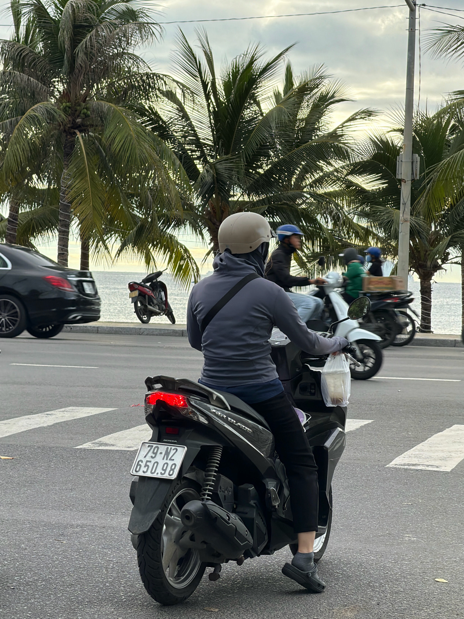 This image depicts a street scene in Nha Trang, Vietnam, with a focus on a person riding a scooter. The scooter is black, with a visible license plate reading "79-N2 650.98". The rider is wearing a grey long-sleeved shirt, dark pants, and a light-colored helmet, along with grey socks and black shoes. They are holding a plastic bag with a drink inside in their right hand. The scooter has its brake light illuminated.
In the background, a black car is seen on the left side of the road, and several other scooters are visible further down the street, with riders also wearing helmets. Palm trees line the street, and the ocean is visible in the distance beyond a walkway. The sky is overcast. The road is paved and marked with white pedestrian crossing lines. The overall impression is of a typical street view in a coastal Vietnamese city.