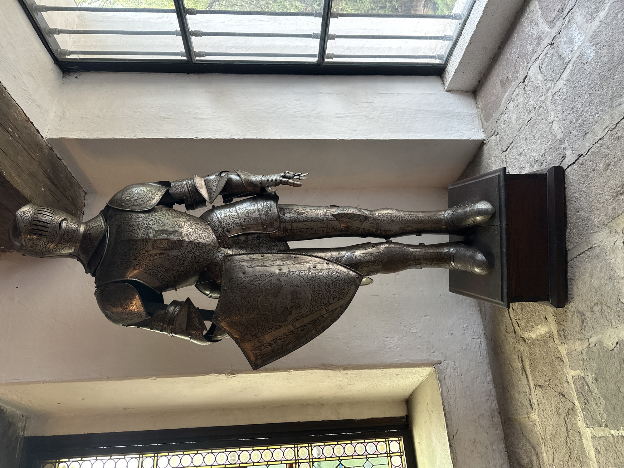 The image shows a full suit of ornate medieval armor displayed in an interior setting. The armor is silver with intricate etched designs. It stands upright on a dark wooden plinth. The background reveals stone walls with plaster trim and windows. The windows are adorned with decorative metal grilles and stained-glass accents. The location appears to be indoors, possibly a museum or historical building, but there are no people visible. The city of Tlalnepantla, Mexico is not apparent from the photograph.