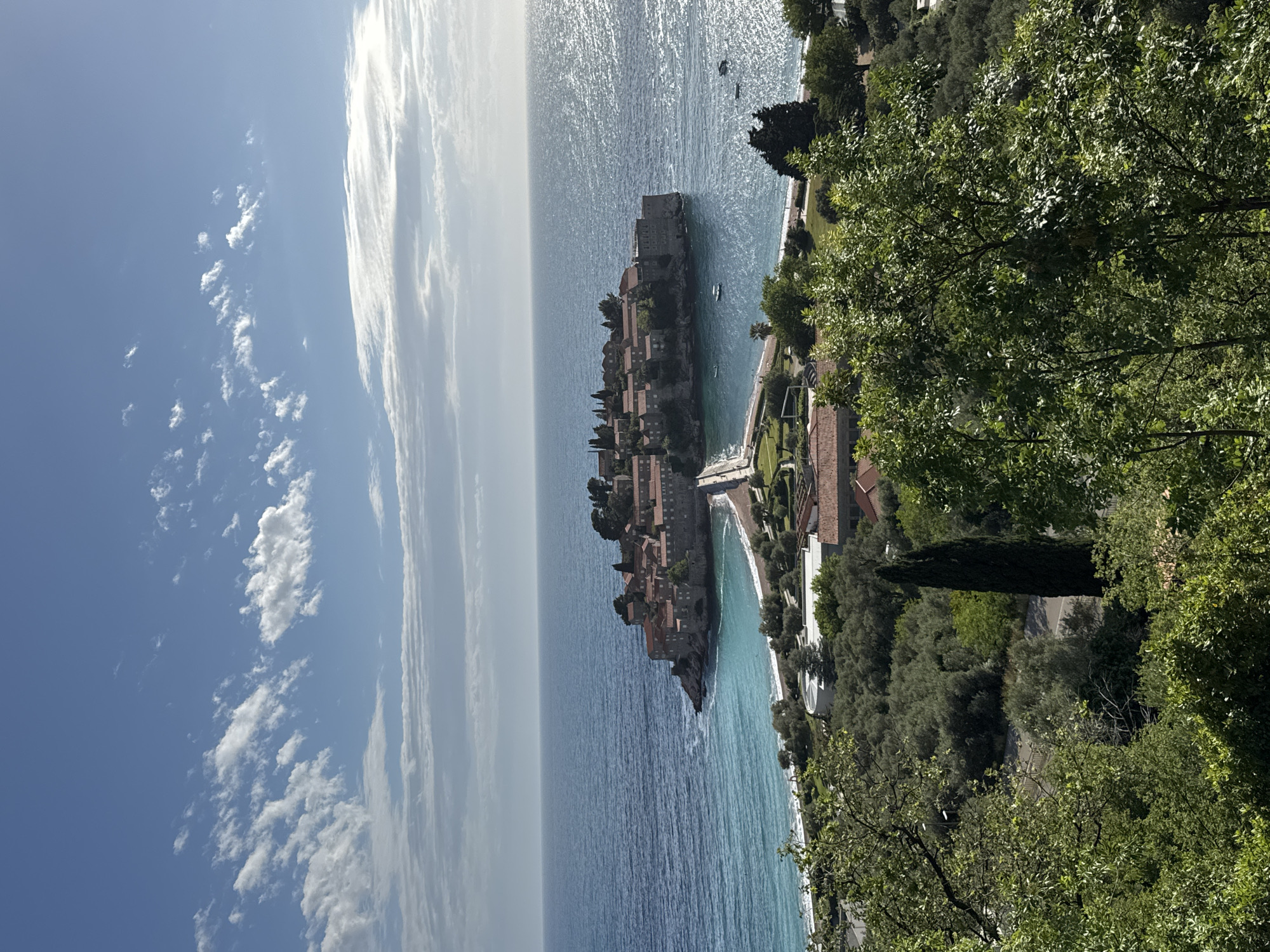 Captured from an elevated vantage point, the landscape exhibits Sveti Stefan, Montenegro. The island, densely populated with buildings featuring terracotta roofs, is attached to the mainland by a narrow isthmus. Calm, azure water surrounds the island. To the right of the island, a coastline extends, fringed by a beach. Inland, lush vegetation and trees thrive along the coast. The scene transitions from the sea to a bright blue sky with scattered clouds.