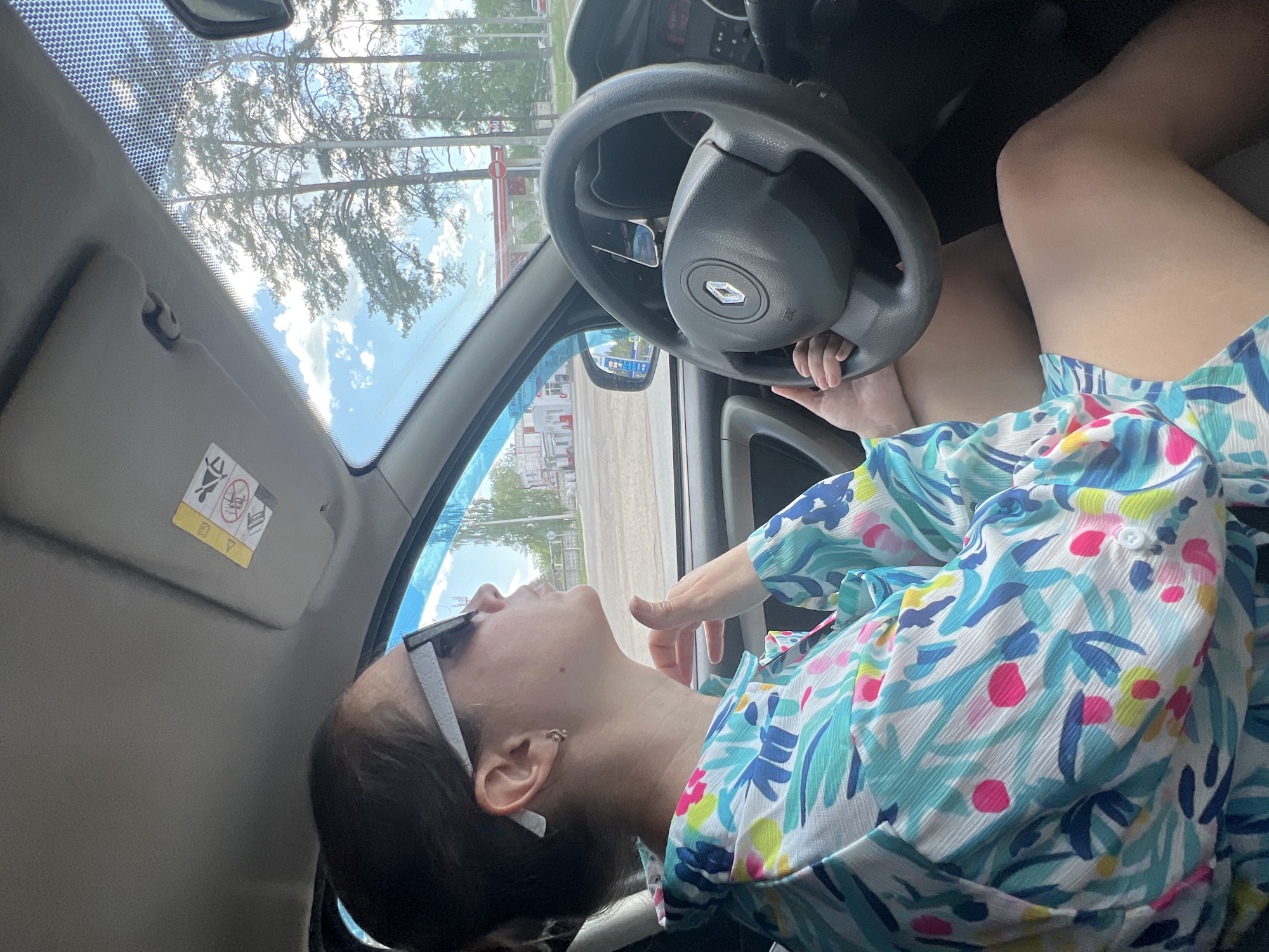 A woman with dark hair and sunglasses resting on her head reclines in the driver's seat of a car. She is wearing a floral-patterned shirt. Her right hand rests near the steering wheel, which bears a Renault logo. Through the windshield, a street scene is visible, with trees and a building with red and white accents. This location is in the city of Chaikovsky, Russia.