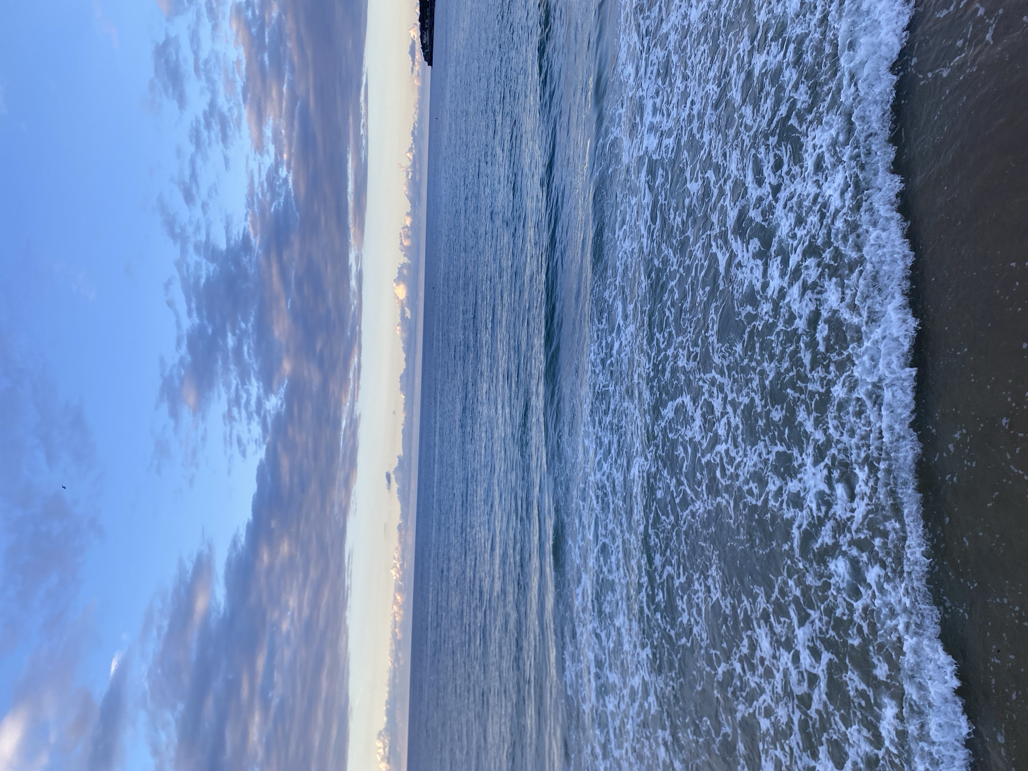 The image captures a seascape under a sky with layered clouds, likely taken in Gijón/Xixón, Spain. The view is a coastline, the left side of the frame showing the sky with a mix of blue and cloudy formations, some tinged with hues of sunset. Adjacent to the sky is the ocean, transitioning from relatively calm waters in the center to frothy waves breaking near the shore on the right edge. The details of the land are not visible, focusing on the sea and sky interface. There are no people visible.