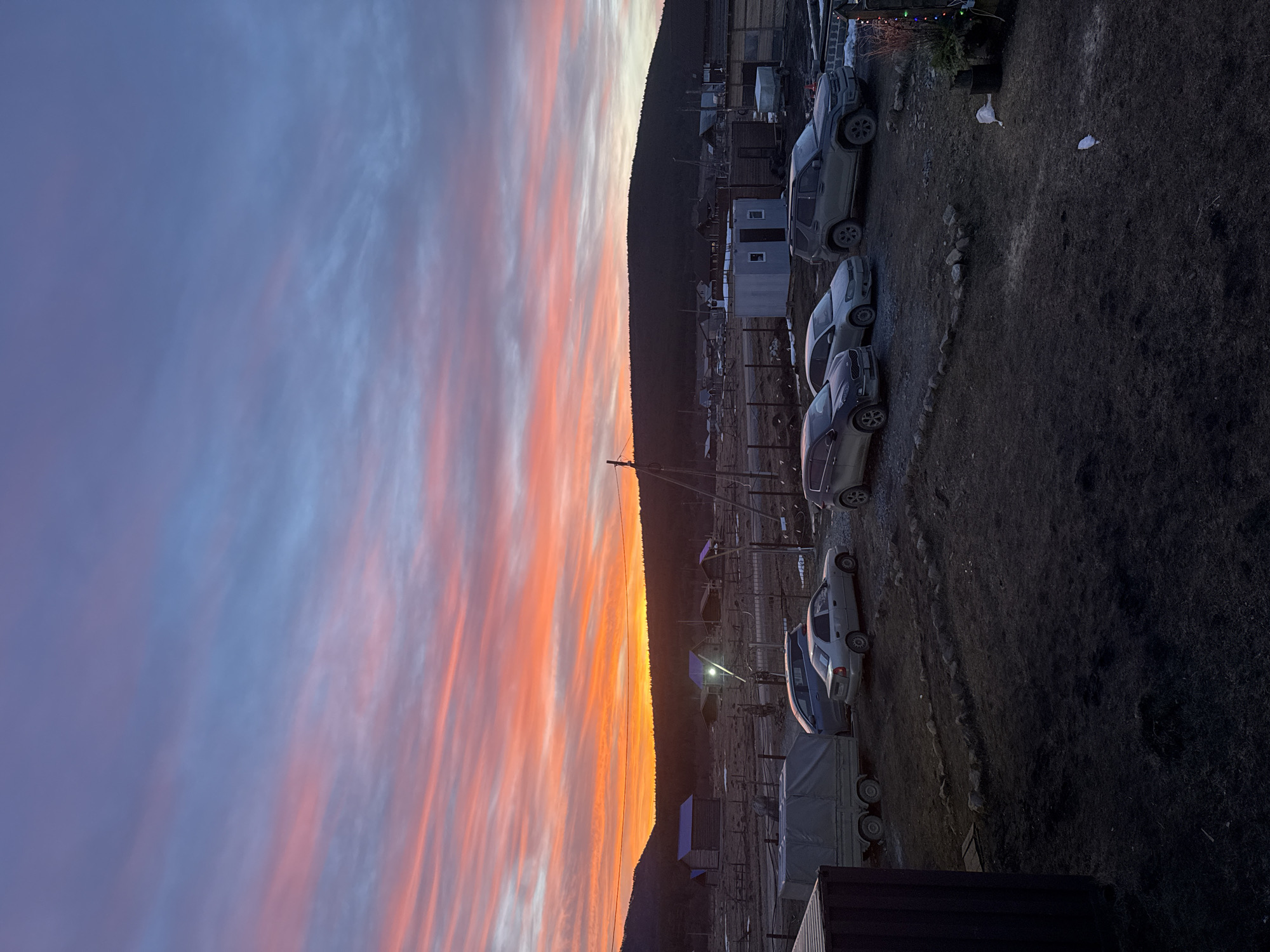 This image captures an outdoor scene in the city of Tyulyuk, Russia, at either sunrise or sunset. The sky exhibits horizontal bands of intense orange and pink clouds along the horizon, transitioning to lighter blue and purple hues in the upper atmosphere. A dark, forested mountain range forms the distant backdrop.
The mid-ground features multiple small, rustic wooden structures, some with blue roofs, dispersed across uneven, dark ground. A bright light source is visible from one of these structures. Fences and utility poles with overhead wires crisscross the area. A small, light-colored corrugated metal shed with a dark door is visible to the right of the center.
In the foreground and mid-ground, six vehicles are parked on dark, dirt ground with visible tire tracks. From left to right, these include a grey enclosed utility trailer, followed by a dark grey sedan, two lighter-colored sedans, a silver/grey SUV, and a partial view of another dark vehicle on the far right. All visible vehicles appear soiled with dirt or dust.
The ground is predominantly dark earth, with some sparse, dry vegetation and minimal patches of snow or ice. A line of small rocks delineates a section of the foreground ground. No individuals or animals are present. A white plastic item and a dark bucket are visible on the ground in the right foreground, near some dried plants with decorative string lights.