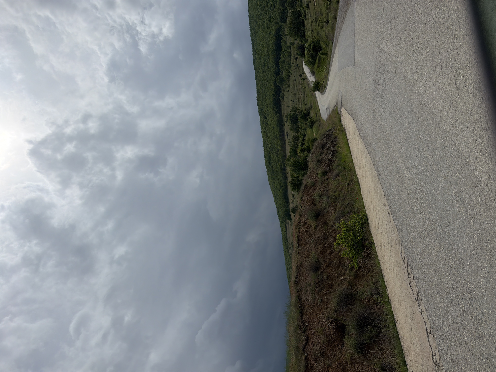 The image shows a paved road winding along a hillside near Drenkovo, Bulgaria. The road surface appears to be asphalt or concrete. The terrain on one side of the road is covered with vegetation, including trees and shrubs. On the opposite side, the sky is overcast with heavy gray clouds, suggesting an imminent weather event. The photograph appears to be taken from a low angle, close to the road surface. There are no visible people, animals, or vehicles.