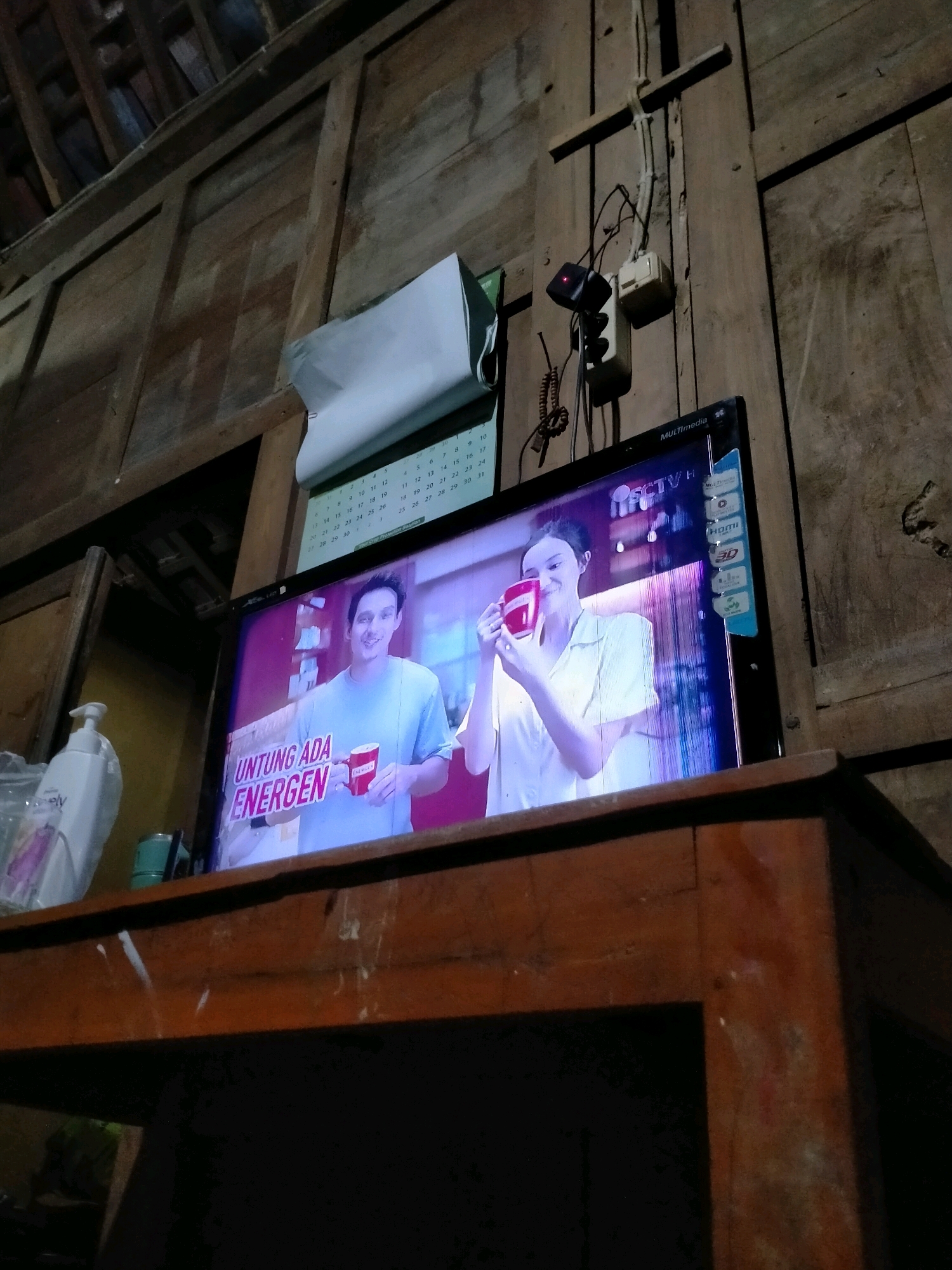 The image depicts a television screen displaying an advertisement for "ENERGEN" in what appears to be a dimly lit, rustic room. The advertisement features two individuals, a man and a woman, both holding red mugs and smiling. The man is wearing a light blue t-shirt and the woman is wearing a white shirt. Text on the screen reads "UNTUNG ADA ENERGEN". Above the television, a calendar is visible, with some days circled. Electrical wires and a power outlet are mounted on a wooden wall behind the television. To the left of the television, a white pump bottle, possibly hand sanitizer or soap, is placed on a wooden surface. The background suggests an interior with wooden walls and ceiling. The overall impression is of a domestic setting, likely a living room in Playen, Indonesia, where a television is being watched.
