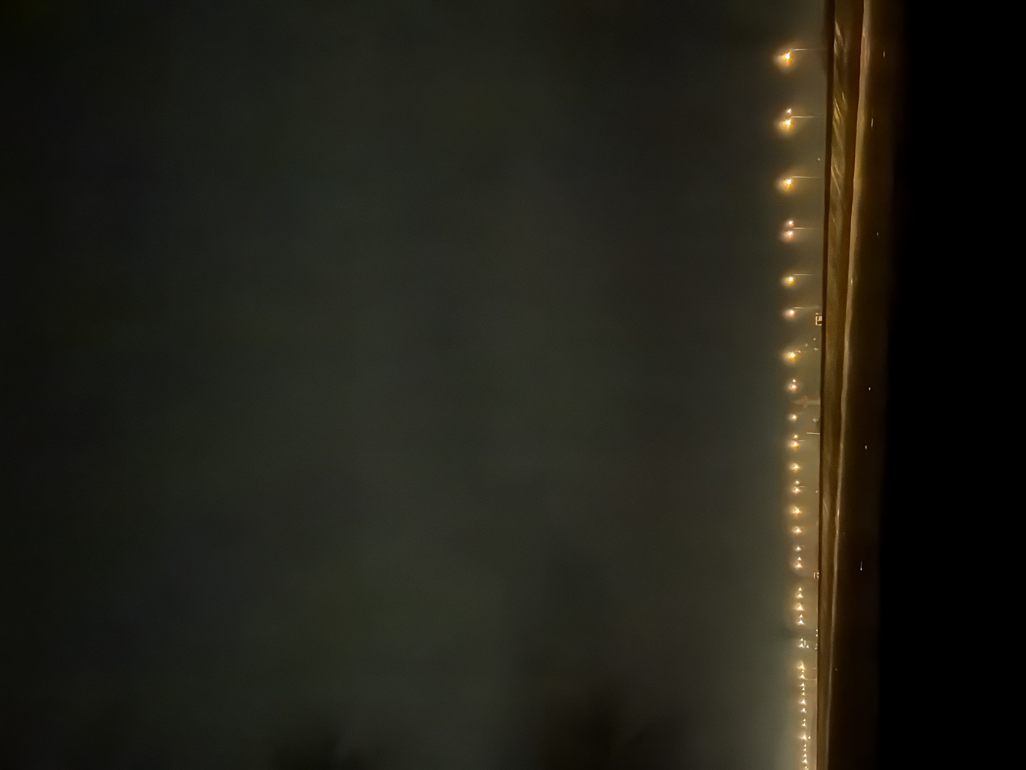 The image shows a night scene along a road or bridge in Noviy, Russia. A row of evenly spaced, warm-toned streetlights stretches into the distance along the right side. The left side of the image shows a dark, hazy expanse, possibly indicating fog or low clouds. A dark structure, likely a barrier or wall, runs vertically along the right edge of the image. The overall lighting is dim, and the scene appears to be quiet and still.