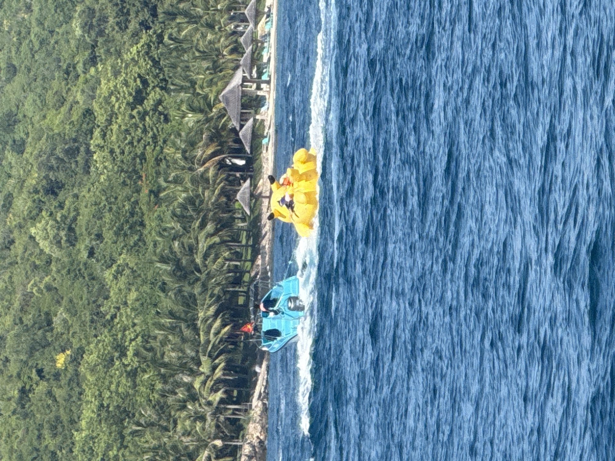 In the city of Nha Trang, Vietnam, two vessels are navigating the blue ocean waters off the coast of a lush, green island covered in palm trees. In the foreground, a bright yellow inflatable tube, shaped like the character Pikachu, is being towed by an unseen boat, creating a white wake behind it. A person is visible riding on the Pikachu inflatable. To the left, a small, blue motorboat with a canopy is moving through the water, also creating a wake. A person is steering the blue boat. In the background, the island shoreline is visible with a sandy beach dotted with thatched-roof cabanas and lounge chairs. The island is densely forested with various shades of green foliage, including many palm trees.