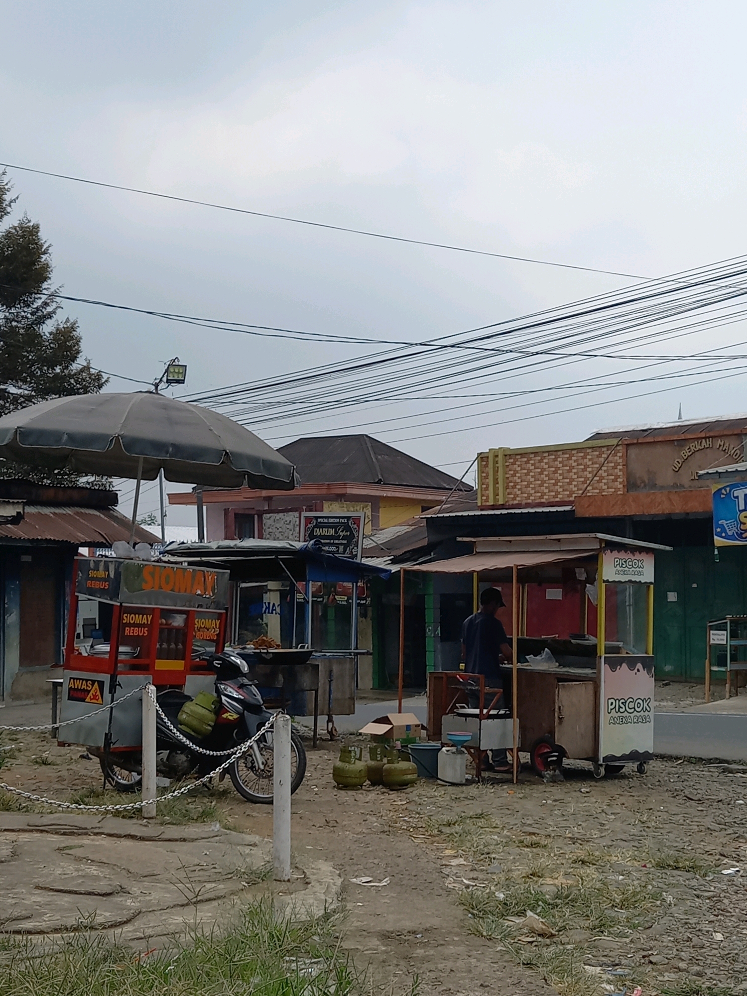 The image depicts an outdoor street scene in Sangubanyu, Indonesia, likely near a road given the overhead power lines and the presence of multiple food stalls. In the foreground, a red and yellow food cart, identified by "SIOMAY REBUS" signage, is parked with a black motorcycle partially attached to it. The motorcycle has a green gas cylinder strapped to its rear. Several other food carts are visible, including one with "PISCOK ANEKA RASA" branding and another with a blue canopy. One person, wearing a dark blue shirt and a black cap, is visible standing behind the "PISCOK" cart, appearing to be tending to it. Two green gas cylinders are placed on the ground near this cart. The environment is dusty and unpaved, with patches of dry grass and some concrete elements. Several buildings are visible in the background, including structures with corrugated metal roofs and a more modern facade with brick-patterned accents. The sky is overcast.