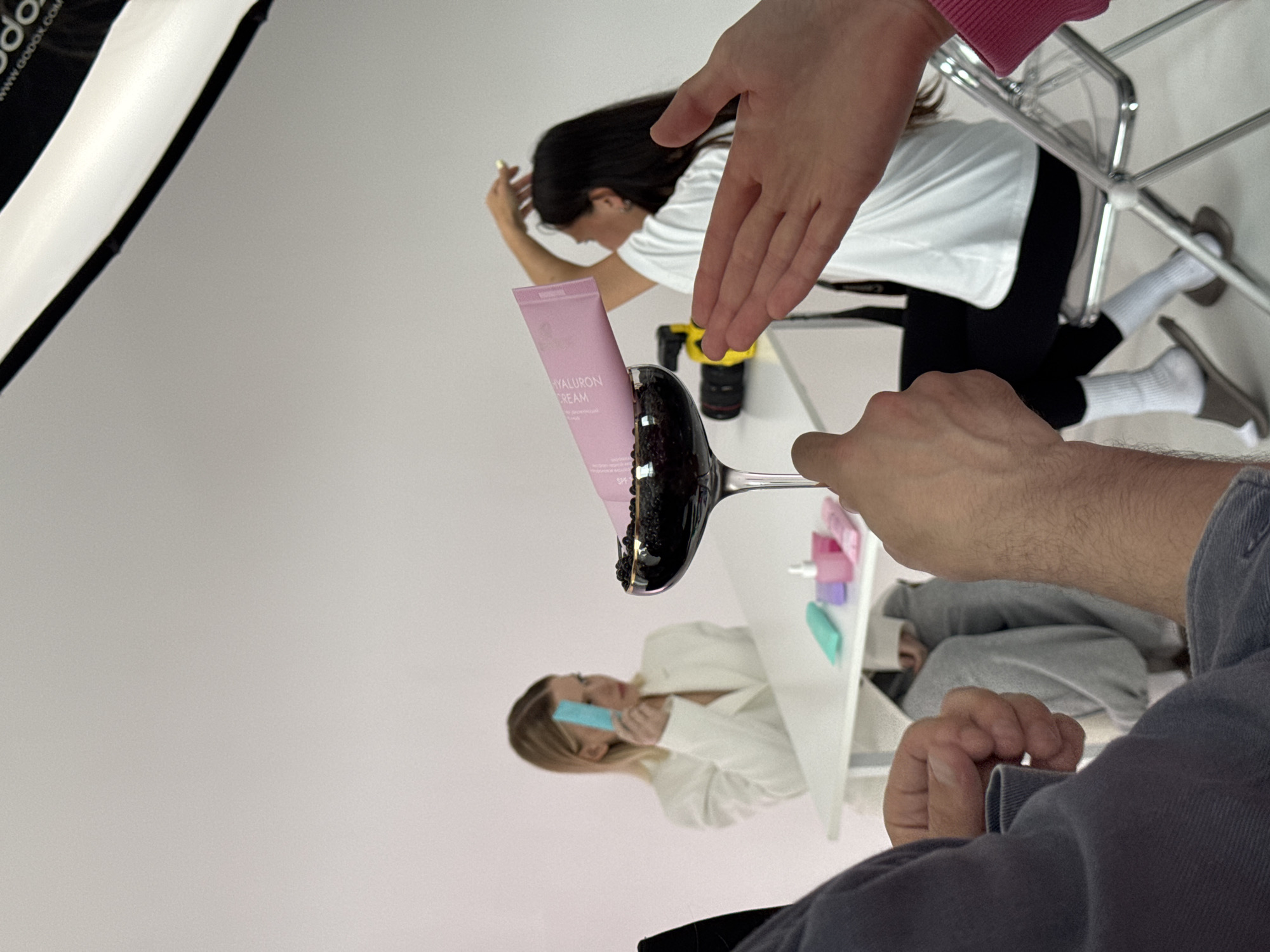 The image appears to be a behind-the-scenes shot of a photoshoot in a white studio. A pink tube of "HYALURON CREAM" is placed upright in a wine glass filled with black objects resembling beads or caviar. Multiple hands are visible in the frame, suggesting adjustments or staging of the product. Two models are present; one is seated in the background wearing a white t-shirt and black pants, and the other is at a white table wearing a white blazer. Various makeup items and tools are on the table. Lighting equipment is visible in the upper left corner. There is no indication of Sochi, Russia.