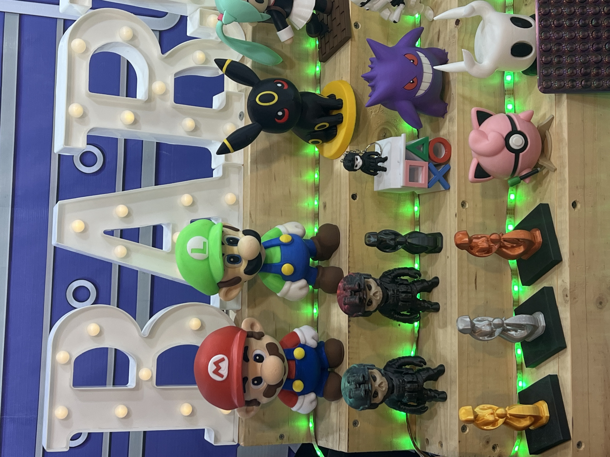 This media file captures a vibrant display of pop culture and gaming-themed figurines and decorative items, likely arranged on a wooden wall or display board. The background consists of vertical wooden planks, illuminated by a string of green LED lights that run along the seams. To the left, large white illuminated letters, possibly spelling out "BAR," are mounted against a blue panel with grey vertical stripes.
Numerous figurines are visible, including prominent representations of Mario and Luigi from the Super Mario Bros. franchise, positioned near the illuminated letters. Mario wears his classic red hat with an 'M', and Luigi sports his green hat with an 'L'.
Other characters include several Pokémon figures: a black Umbreon with yellow rings on a yellow base, a purple Gengar with a wide grin, and a pink, round Jigglypuff-like character with a black mask detail and what appears to be a microphone. A white, ghost-like figure, resembling the protagonist from Hollow Knight, is also present.
Further elements of the display include several dark, skull-faced, armored figures, some resembling Call of Duty characters, standing on black bases. Three distinct trophy-like statuettes, colored gold, silver, and copper, are arranged on black pedestals in the lower right. A small keychain with a black skull figure hangs near the Umbreon. Partially visible in the top left is a light green figure, possibly Hatsune Miku. A small white block displaying the iconic PlayStation controller symbols (triangle, circle, X, square) is also part of the setup. A purple, textured item, resembling a pop-it fidget toy, is partially visible in the top right corner.
The display is brightly lit, with the green LEDs and the internal lights of the large letters providing a decorative glow. The arrangement suggests a collection, merchandise display, or decoration for a game room or event.