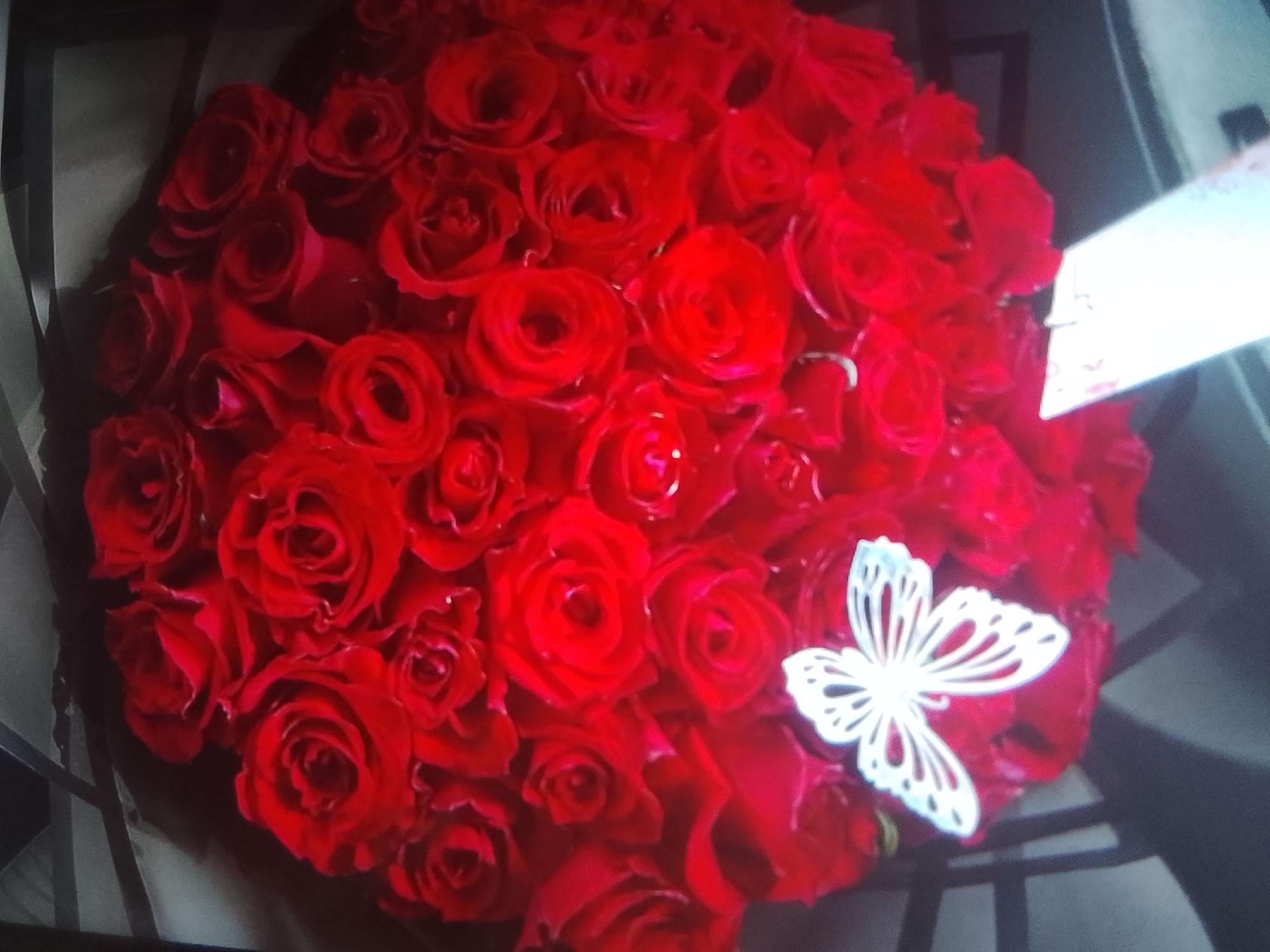 The image displays a close-up, angled view of a large bouquet of vibrant red roses. The roses are densely packed and appear to be fresh, with many showing fully opened petals and deep red hues. In the center of the bouquet, a white, decorative butterfly is placed, its intricate cut-out design contrasting with the rich color of the flowers. The background is dark and appears to be out of focus, suggesting the lighting conditions or the depth of field of the photograph. The composition is dominated by the textures and colors of the roses. There is no indication of a specific location within Howayi, Nigeria, or any people, actions, or interactions visible in the image.