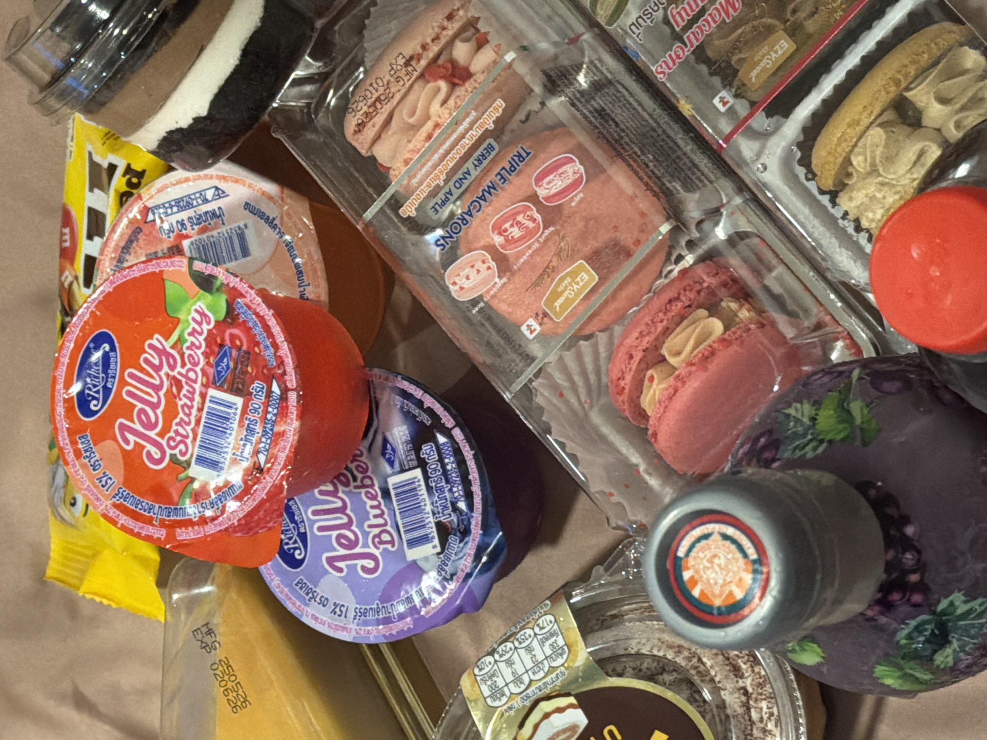 The image shows an overhead shot of a collection of packaged food items. There are several plastic containers holding dessert items: a multi-layered chocolate and cream dessert, a package labeled "TRIPLE MACARONS" with berry and apple flavors, macarons, and another set of what appears to be cookies.
Additionally, there are two containers of "Jelly" in Strawberry and Blueberry flavors from the brand "Riche". A yellow package lies partially visible beneath one of the jelly containers. The packaging on some of the items has writing in what appears to be the Thai language.
Based on the "70-1-09235" number appearing on the Jelly packaging, and that food is sold in the city of Kathu, Thailand, and is likely being sold in Kathu.