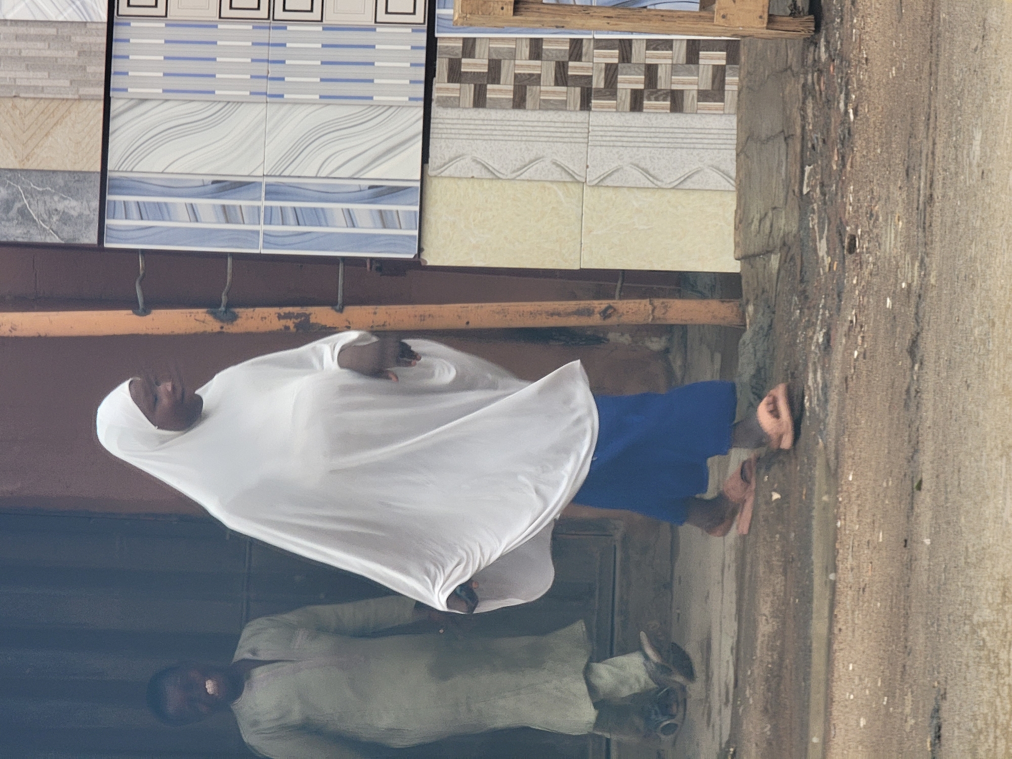 A woman wearing a white hijab and blue clothing stands near a building displaying tiles for sale. A reflection in a window shows a man in light-colored clothing. The location is Maiburiji, Nigeria, as indicated by the prompt.