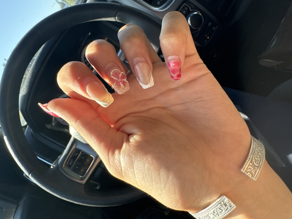 A human right hand, appearing to be female, is prominently featured within the interior of a vehicle. The hand displays a detailed manicure: the thumb and pinky nails feature a marbled pink and red pattern, while the index, middle, and ring fingers have clear/nude bases with white French tips. The middle finger is further adorned with a three-dimensional pink and white floral design. A silver-colored, open-cuff bracelet with embossed scrollwork is worn on the wrist. The hand is positioned over a black steering wheel, which has integrated media or cruise control buttons on its left spoke. Direct sunlight illuminates the hand, casting distinct shadows. Through the vehicle's windshield, a bright blue sky and indistinct green foliage are visible. The specific location within Rancho Cordova, United States, cannot be determined from the provided visual evidence.
