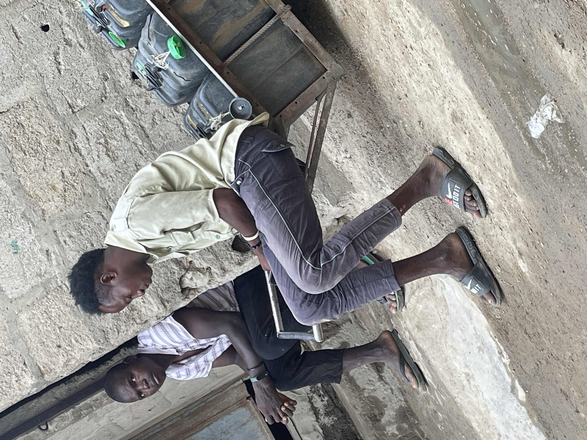 Two men are sitting outside a building. One man is wearing a light-colored shirt and gray pants and is sitting on a metal cart with several containers. The other man is wearing a striped shirt and dark pants. Both men are wearing sandals. The ground is concrete. The location is Unknown, Unknown.