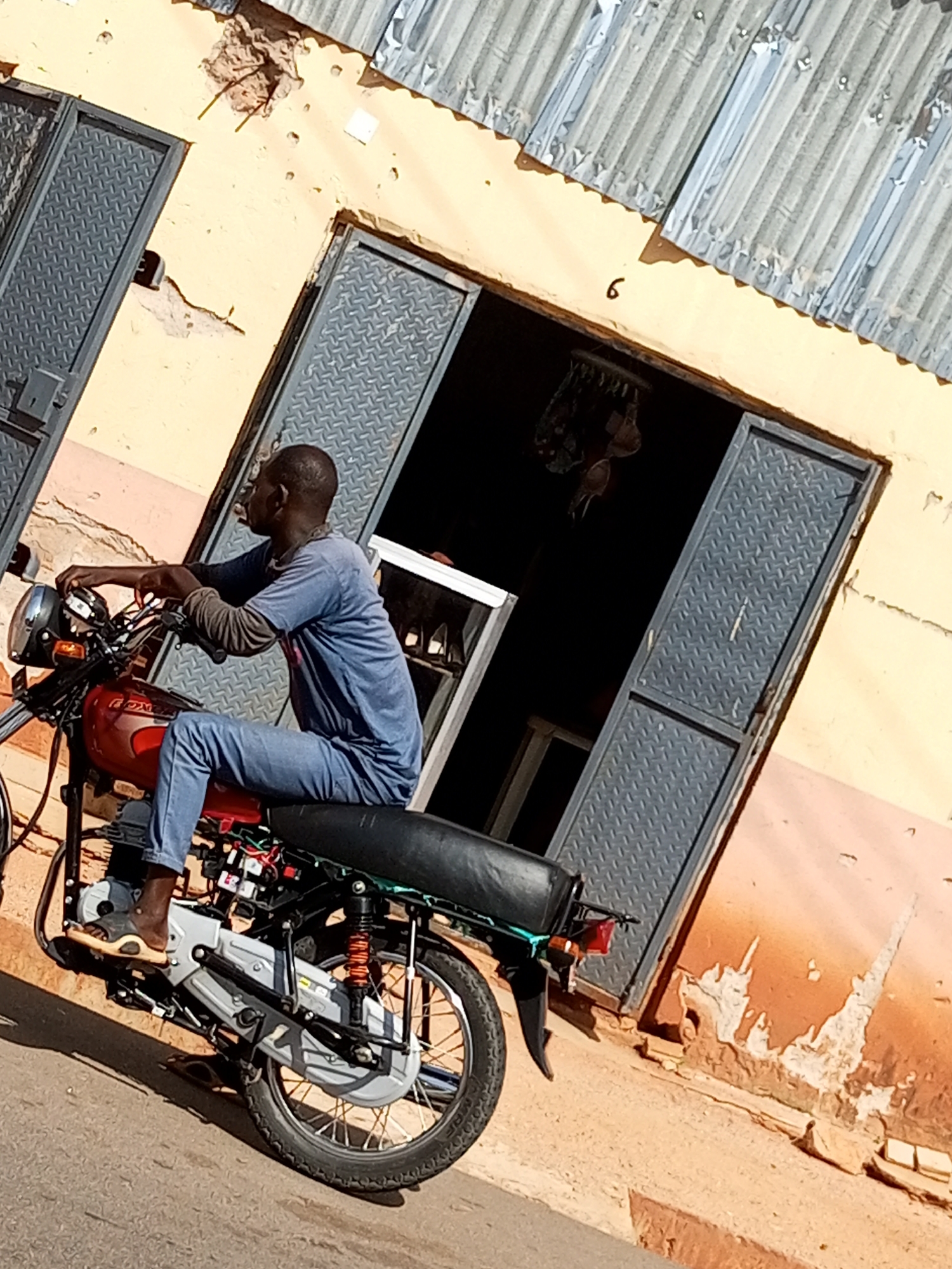 A man is sitting on a red motorcycle in front of a building with a corrugated metal roof. The building has a light yellow facade and two grey metal doors. One door is partially open, revealing a dark interior. The man is wearing a blue long-sleeved shirt and blue jeans. He is looking to the left. The motorcycle has a black seat and a visible engine. The ground is paved. The image appears to be taken during the day, with sunlight casting shadows. The building shows signs of wear and tear, with some areas of peeling paint. A numeral '6' is visible on the facade of the building, near the roofline. This scene is located in Gombe, Nigeria.