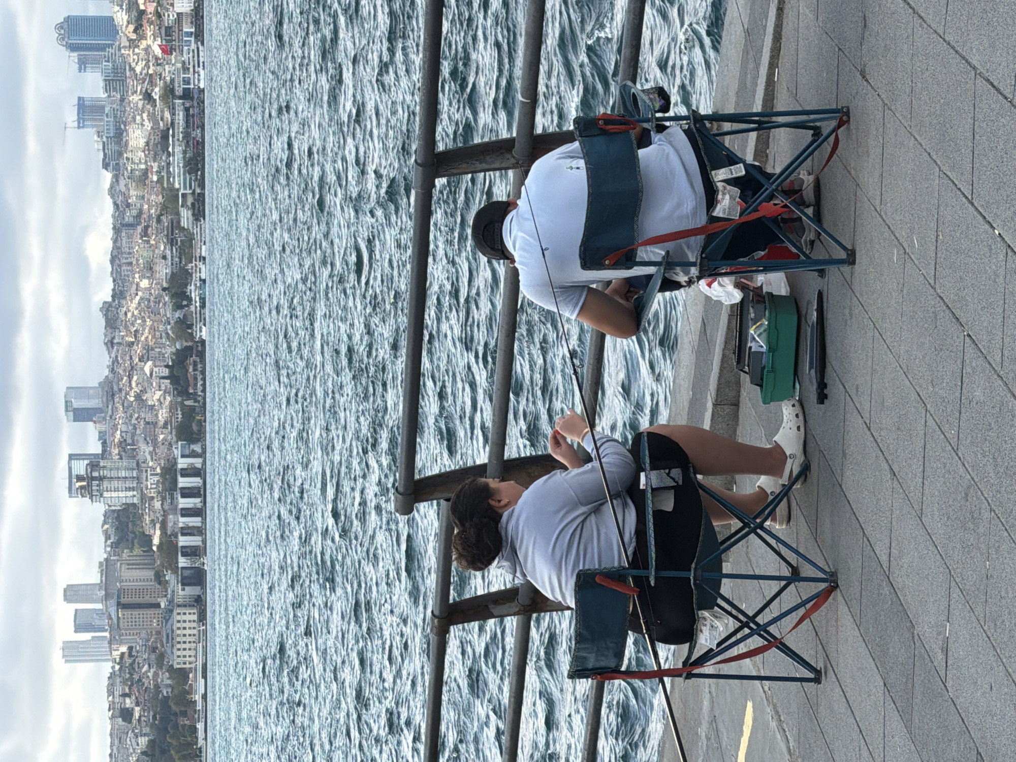 The image shows two people fishing from a promenade overlooking a body of water, with the city of Üsküdar, Turkey, visible in the background.
In the foreground, a woman is seated in a folding chair, facing the water, with a fishing rod resting in front of her. She is wearing a grey hoodie and black shorts. Next to her, a man is also seated in a folding chair, facing the water, with a fishing rod. He is wearing a light blue polo shirt and a dark baseball cap. A green tackle box is on the ground between them, and a fishing rod lies on the ground to the left of the woman's chair. The promenade is paved with large, rectangular grey tiles. A dark metal railing runs along the edge of the promenade, separating it from the water.
The water is a deep blue-green with visible ripples, suggesting a light breeze. In the background, the cityscape of Üsküdar rises from the shore. Numerous buildings of varying heights and architectural styles are visible, including several modern skyscrapers and older, more traditional structures. The sky is overcast with a mix of grey and white clouds.
