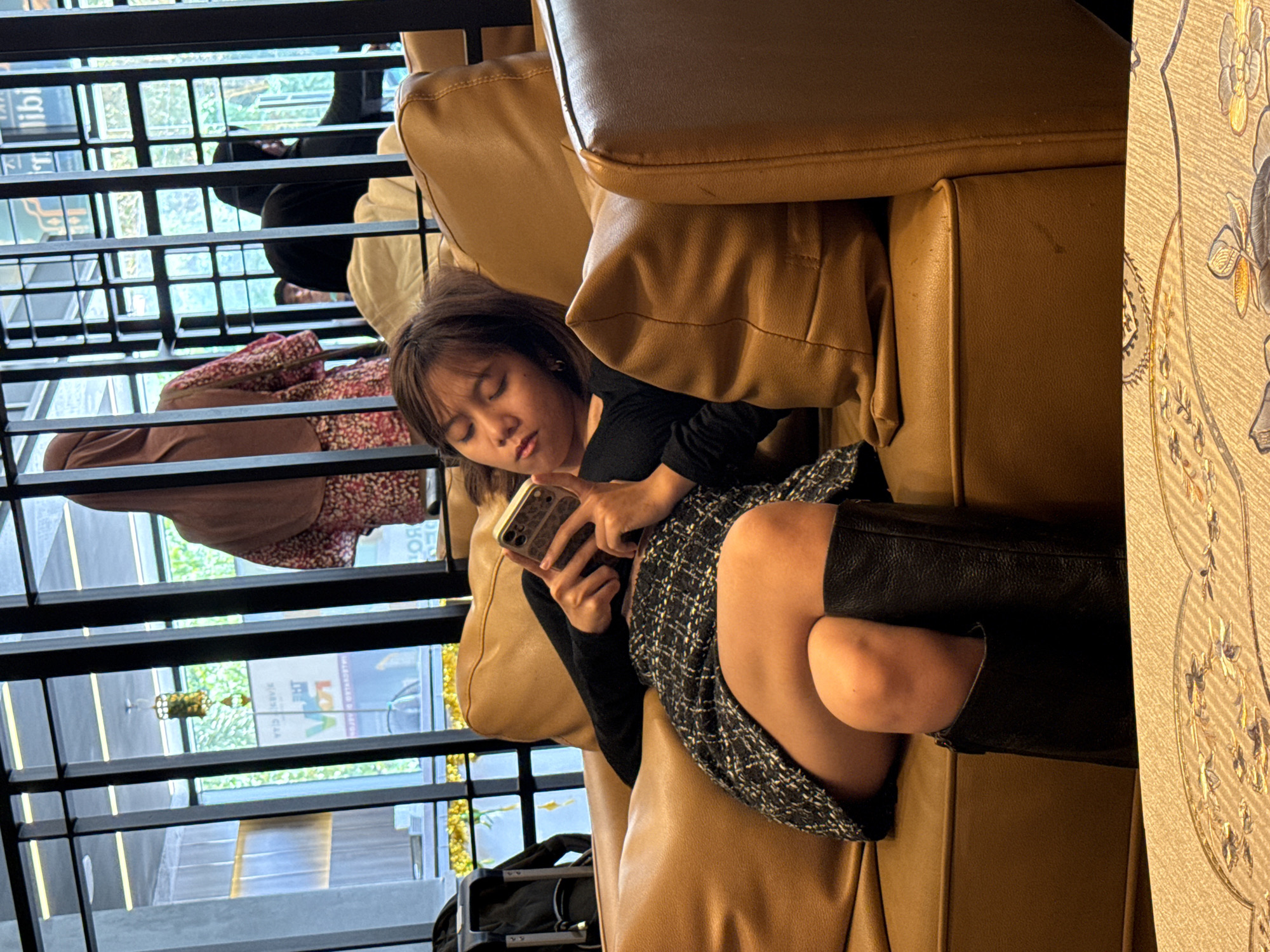A woman sits on a light brown leather couch with a dark tweed skirt, black shirt, and black leather boots. She is holding a phone with a brown and beige patterned case. Behind her, a woman in a floral dress and a head covering sits. Behind them, a black frame appears to be part of a window or wall structure, through which a building with a store called "Loka" is visible. The woman is in Kuala Lumpur, Malaysia. Luggage is on the floor near her feet.