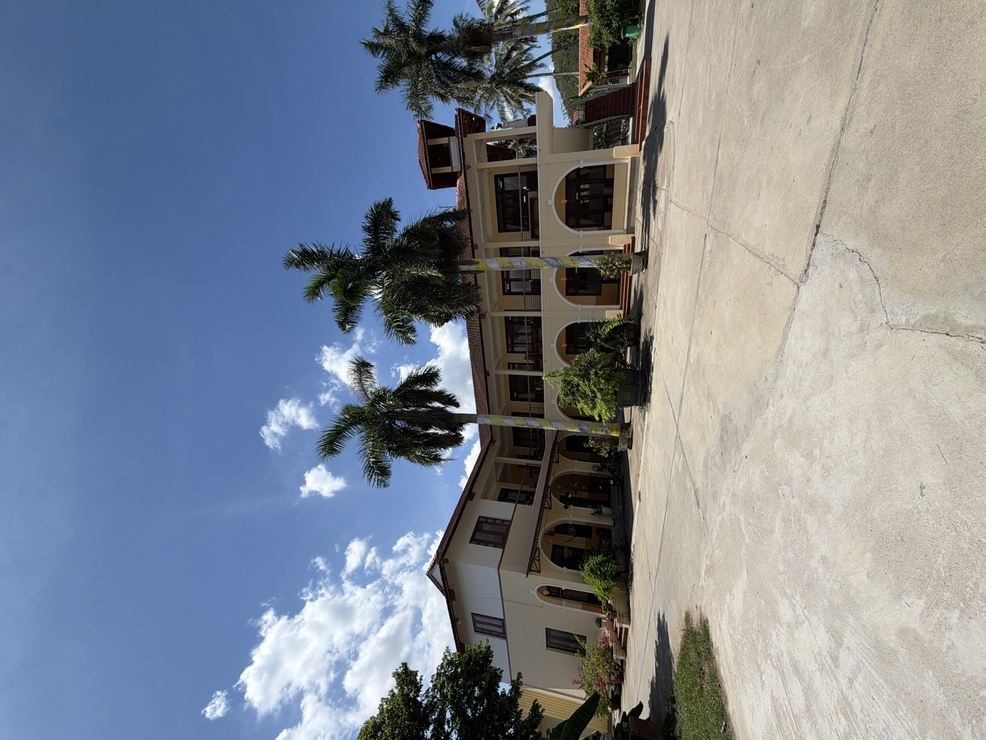 The image shows a two-story building with a light yellow facade and a red tile roof, situated in Hội Tín, Vietnam. The building has arched openings on the ground floor and balconies on the second floor, supported by pillars wrapped in decorative yellow and white fabric. Several tall palm trees are present on the grounds, with lush green foliage. The area in front of the building is paved with concrete, and there are some potted plants and small trees scattered around. The sky is a clear blue with scattered white clouds. The scene appears to be an outdoor common area, possibly a hotel or a public building, with no people visible. The overall environment suggests a warm, sunny climate typical of Vietnam.
