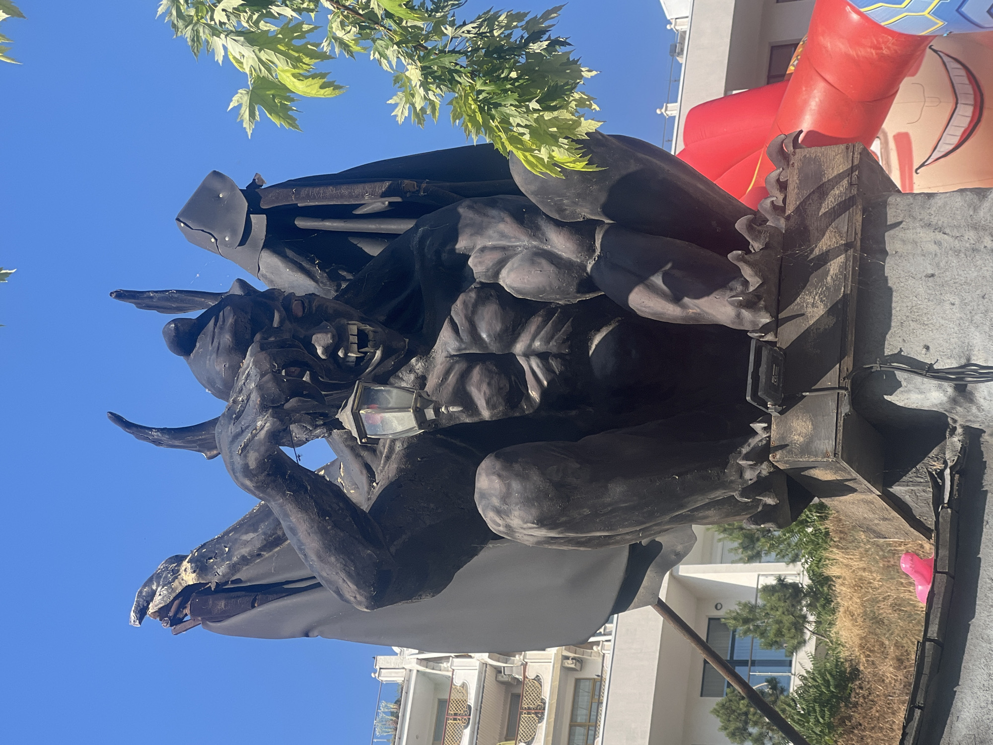 This is a low-angle, vertical, full shot of a dark, gargoyle-like statue with wings, sitting on a wooden beam. The statue is intricately detailed, with a muscular physique, sharp claws, and a menacing face. It appears to be holding a small lantern in its hands. Behind the statue, a large red inflatable object with a cartoonish face can be seen, along with a bright blue sky. Green leaves from a tree hang into the frame from the top left. In the background, buildings with balconies and windows are visible, suggesting an urban setting. The lighting indicates it is daytime, likely sunny. The statue's pose and grim expression convey a sense of brooding or anticipation. The overall impression is one of a macabre or fantasy-themed installation, possibly for an event or decoration in Odesa, Ukraine.
