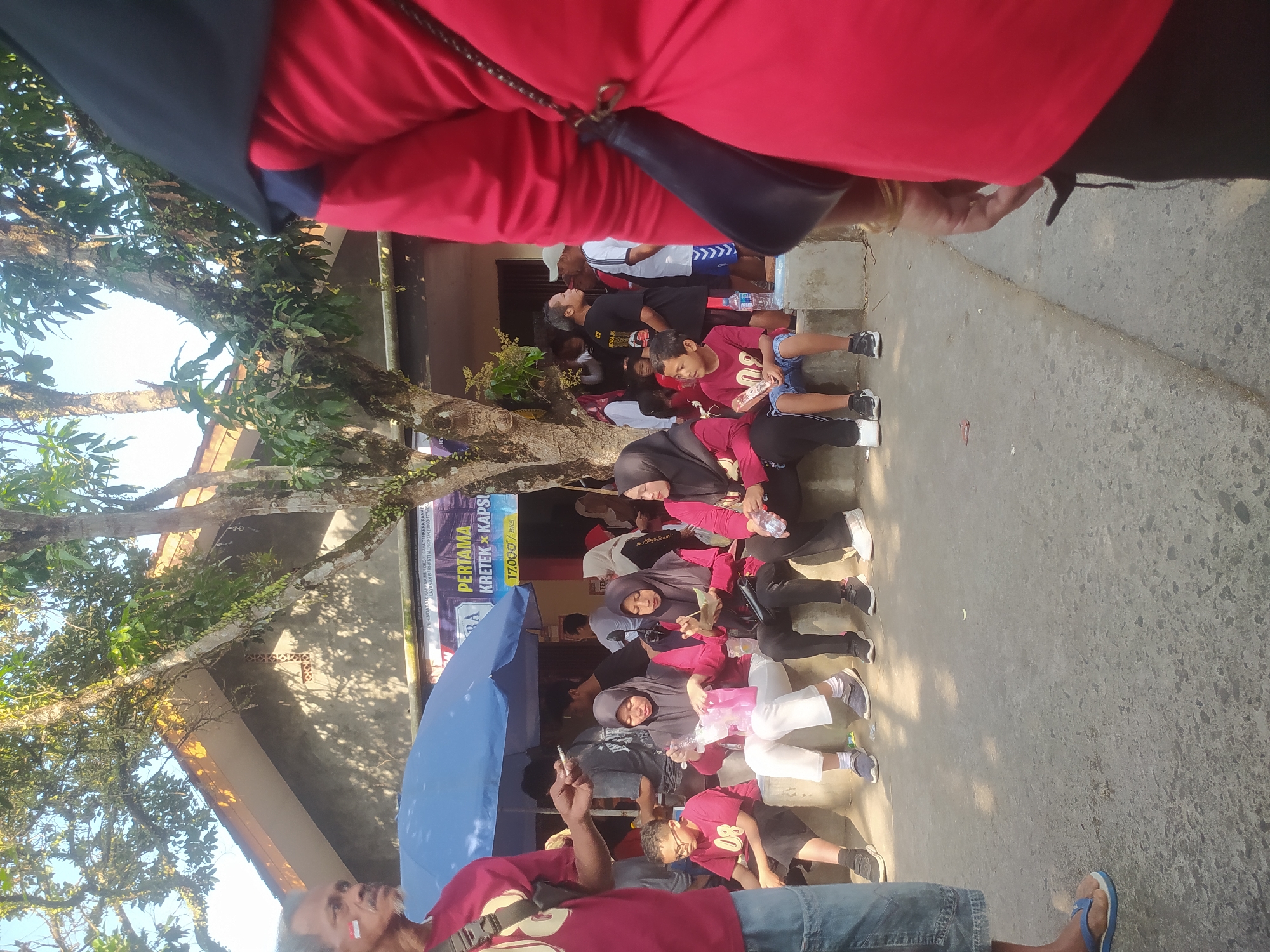 The image depicts a scene in Lopait, Indonesia, likely during a local event or gathering. In the foreground, a person wearing a red shirt and black headscarf is partially visible from the back, with their right arm and hand extended. To the left, a man with a mustache and a red and white mark on his cheek is standing near a blue umbrella. He is wearing a maroon t-shirt and blue shorts, holding what appears to be a cigarette.
Several people are seated on a concrete ledge. Two women in the center are wearing hijabs and red shirts; one is holding a clear plastic bag, and the other is looking at something in her hands. To their right, another woman in a hijab and red shirt is seated. On the left side of the ledge, a young boy with glasses, wearing a maroon t-shirt with the number "08" and blue shorts, is sitting. To his right, another young boy in a red t-shirt and blue shorts is seated, holding a small object.
Behind the seated individuals, other people are visible, some standing under the blue umbrella, including men in t-shirts and one wearing a white shirt with blue stripes. A building with a grey facade and a yellow-tiled roof is in the background. A large tree with branches extends across the top of the image. A banner with Indonesian text, including "PERTAMA KRETEK KAPS", is partially visible behind the umbrella. The ground is paved with concrete. The overall atmosphere appears to be a casual outdoor gathering.