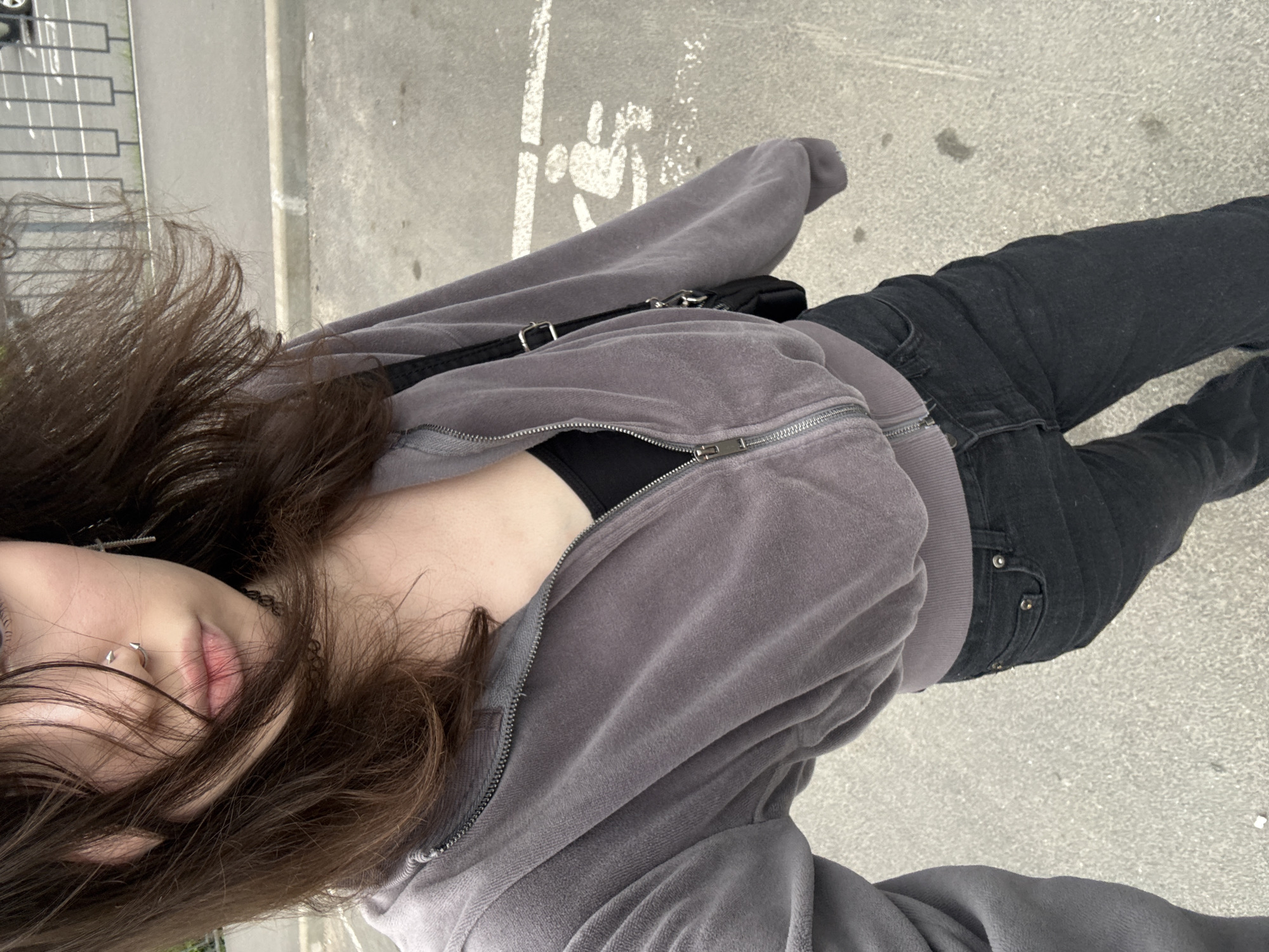A young woman stands on an asphalt surface, potentially a parking lot, given the presence of white painted markings that resemble a wheelchair-accessible parking symbol. She is wearing a grey velour zip-up hoodie over a black tank top and dark wash jeans. Her dark brown hair is windswept, partially obscuring her face. She has a nose ring and a cross earring. A black purse with a silver buckle is slung over her shoulder. The background shows a metal fence and a road.