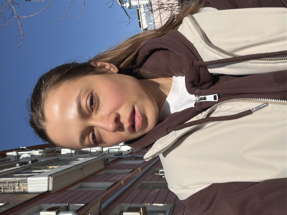The image displays an adult female in a selfie, positioned outdoors on a clear, sunny day in Moscow, Russia. She has medium brown hair, pulled back from her face, which shows strong highlights from direct sunlight. Her lips are slightly parted, and her expression is neutral. She is dressed in a white t-shirt layered under a brown hooded sweatshirt, topped with a light beige puffer vest, all zipped. The background on the left features a multi-story residential building with light red walls, white-framed windows, numerous external air conditioning units, and small balconies, one of which has a satellite dish. The background on the right shows a clear blue sky with bare tree branches and a partial view of another light-colored building.