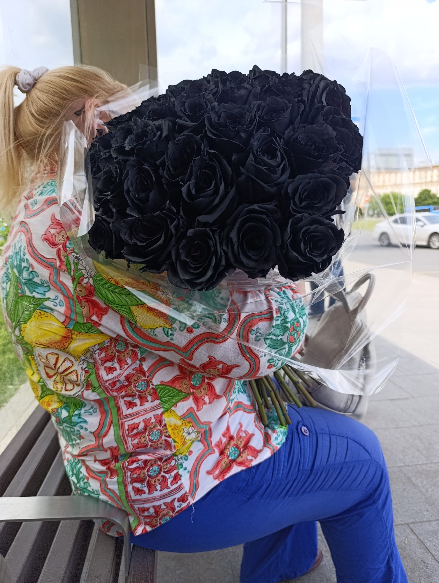 A blonde woman, with her hair styled in a high ponytail and secured with a scrunchie, is seated on a slatted wooden bench outdoors in Moscow, Russia. She is wearing a vibrantly patterned blouse with a white background, featuring red, green, yellow, and blue motifs that include lemons and floral designs, paired with bright royal blue pants.
The woman holds a very large and striking bouquet of black roses, which largely obscures her face. The bouquet is wrapped in clear plastic, emphasizing the unusual, dark hue of the roses. A light-colored handbag is visible behind the flowers, resting beside the woman on the bench.
The scene takes place during the daytime, under what appears to be a clear or partly cloudy sky, with ample natural light. In the background, there are blurred indications of an urban environment, including modern buildings and a street with a white car. Some greenery is also visible to the left of the woman. Her activity involves holding or receiving the bouquet, with her gaze directed downwards towards the flowers. Due to her face being obscured, her emotions are not discernible. There is no visible text in the image.