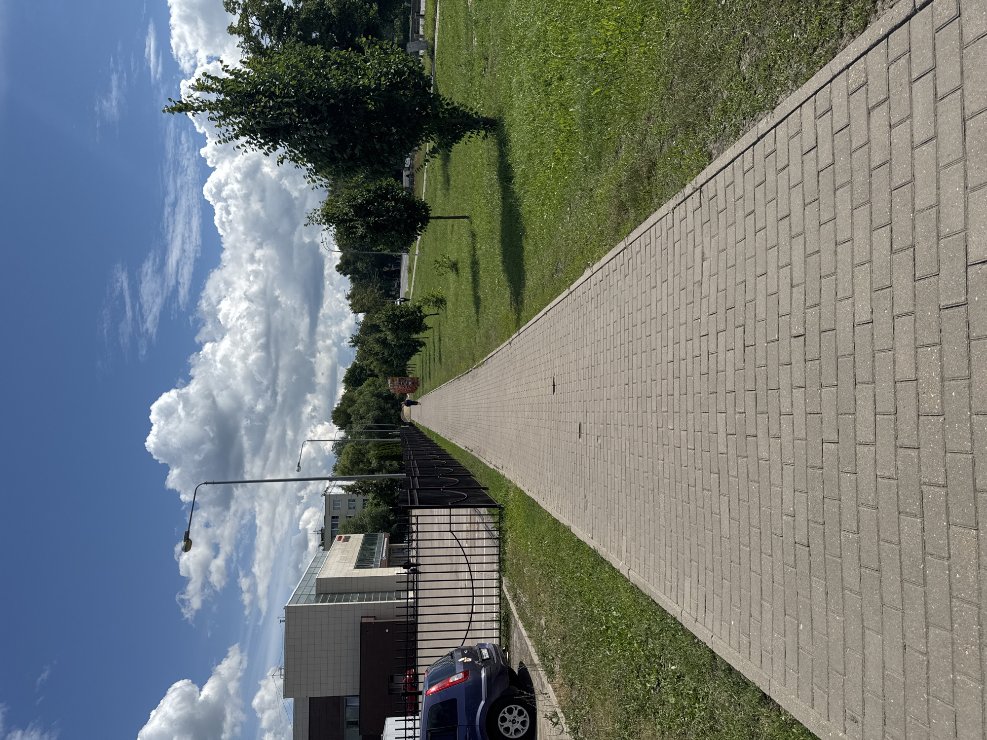A paved walkway made of light-colored rectangular bricks stretches into the distance, bordered by a grassy area on the right and a black metal fence on the left. A blue car is parked to the left of the walkway, near a modern-looking building with a light-colored facade and dark trim. Beyond the fence, there are several streetlights and trees. The sky is blue with scattered white cumulus clouds. A lone figure is visible walking away from the camera down the walkway, and another person is walking further in the distance. The scene appears to be a public space, possibly a park or a commercial area, on a bright, sunny day. This image was taken in the city of Bronnitsy, Russia.