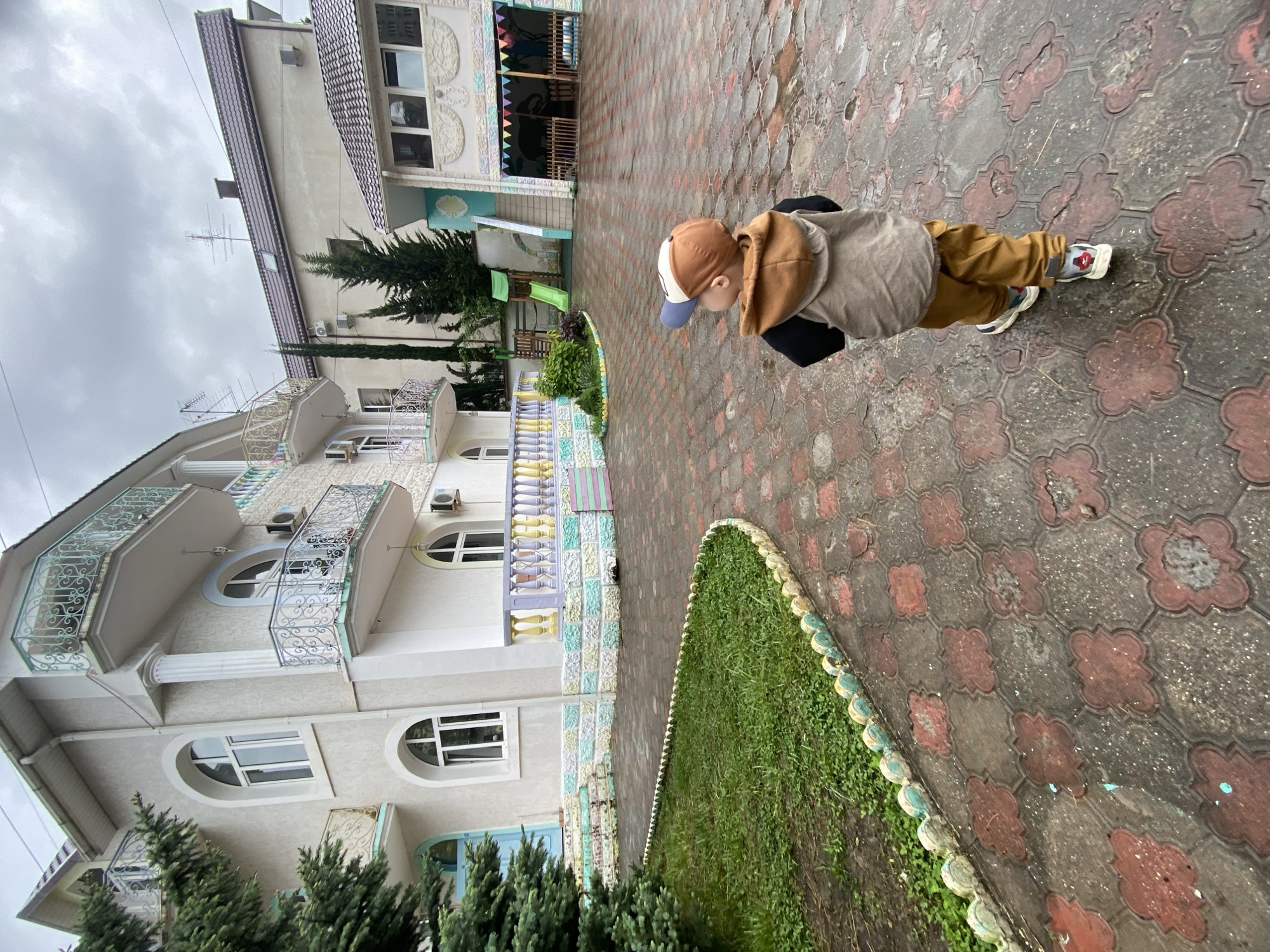 A medium-angle, outdoor shot shows a child standing on a patterned brick walkway next to a green lawn and a large building, possibly a hotel or apartment complex. The child is wearing a brown and white baseball cap, a gray hoodie, brown pants, and sneakers. The child is leaning forward, looking down at something on the ground. The building behind the child is multiple stories tall and has a white facade with arched windows and balconies with ornate railings. The walkway is made of red and gray bricks arranged in a floral pattern. A strip of green lawn with a decorative border runs alongside the walkway. In the background, there is another building with a playground. The sky is overcast. The location is likely in Sochi, Russia, due to the architecture and landscaping style.