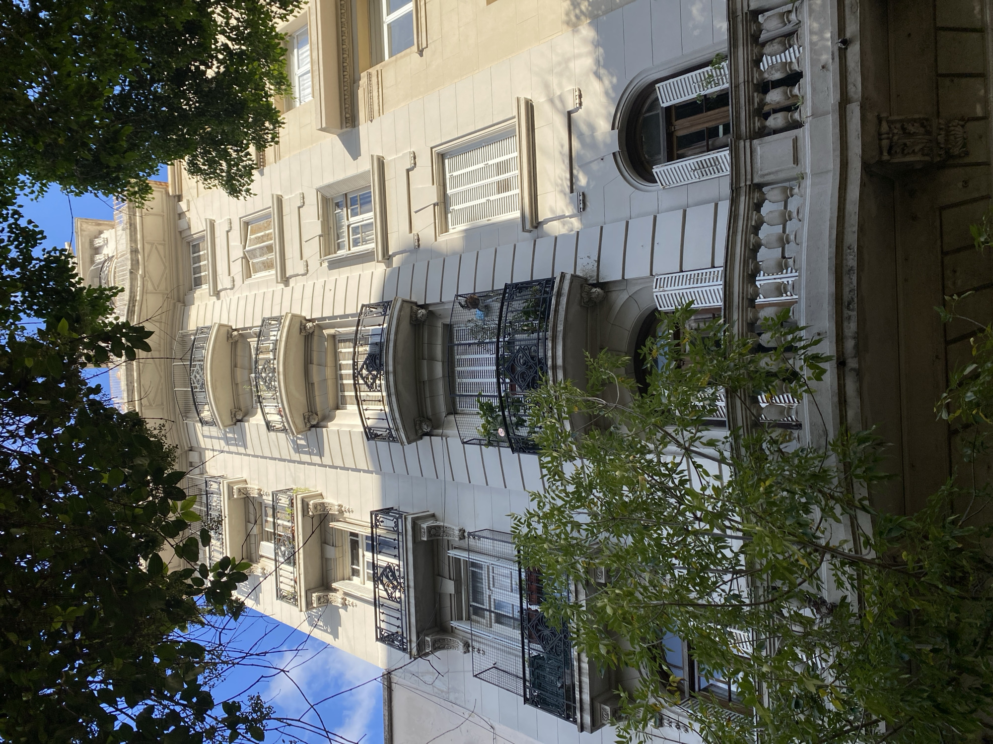 The image depicts a multi-story building facade in Buenos Aires, Argentina. The building is light-colored with ornate architectural details, including projecting balconies with intricate wrought-iron railings. Windows of various shapes and sizes punctuate the facade. Mature trees frame the building on both sides, partially obscuring the view of the blue sky with scattered clouds. The image is taken from a low angle, looking up at the building. There are no people visible in the image. The building's architectural style suggests it may be located in a historic neighborhood within Buenos Aires.