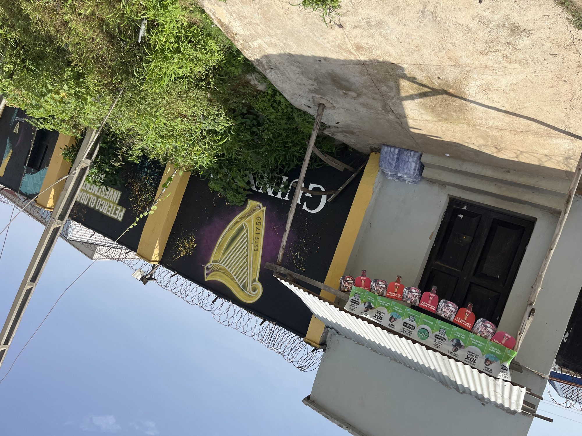 The image captures a street-level view of a building exterior and a surrounding wall in Bukuru, Nigeria. The building features a corrugated metal roof extending over an entrance with a dark-colored door and several steps. Along the edge of the roof, multiple colorful banners are hung, displaying various advertisements, including "Glo" and "Coca-Cola." To the right of the building, a wall is adorned with a large mural depicting the Guinness harp logo and the text "ESTP 1759." Further along the wall, the text "PETERCO GLOBAL INVESTMENT" is visible. The wall is topped with razor wire for security. A collection of bottled water packages is stacked near the building's entrance. Lush green vegetation grows along the base of the wall. A concrete pole with electrical wires extends vertically in the foreground. The sky above is clear and blue.