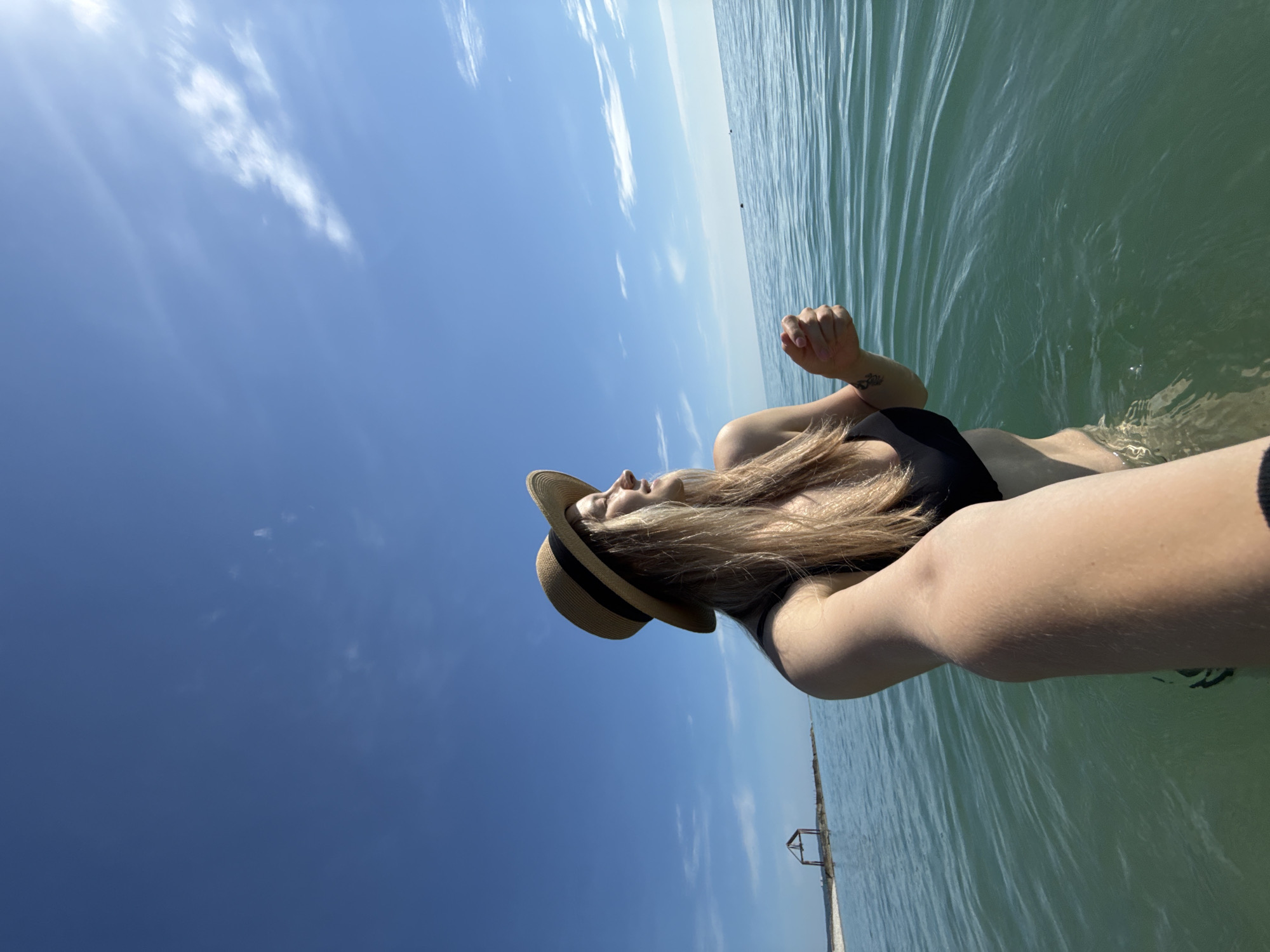 A young woman wearing a black bikini top and a straw hat stands in the shallow, clear turquoise water of a beach. The water is calm with gentle ripples. The sky above is a vibrant blue with scattered white clouds. In the distance, a small, rudimentary wooden structure stands on a sandy spit of land. The woman is facing to her right, with her eyes closed and a slight smile on her face, seemingly enjoying the sun. Her arm is extended forward, holding a phone to take a selfie. The photo is taken from a low angle, giving a sense of being immersed in the water. This scene appears to be at a beach in Nebug, Russia, known for its pebble beaches and clear waters.