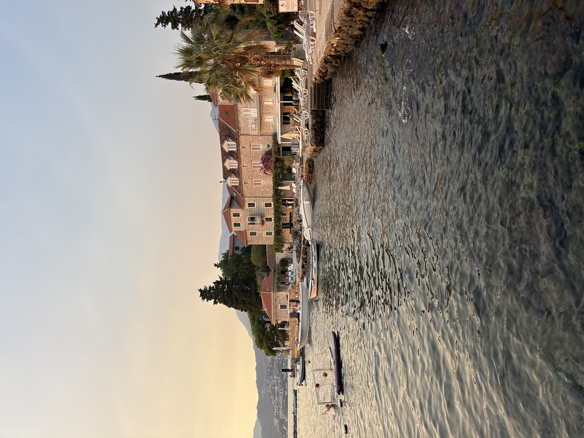 The image depicts a serene waterside scene at dusk in an unknown city. The foreground is dominated by calm, rippling water reflecting the soft, golden light of the setting sun. The water appears clear, with submerged rocks and seabed visible in places. A small, white buoy floats near the left side of the frame.
Further back, a line of several boats is moored near the shore. One of these boats is engaged in a game of water polo, with a person in the water reaching for a ball near a portable goal.
Along the coastline, a row of buildings with traditional architecture, featuring stone facades and red-tiled roofs, lines the waterfront. Some of these buildings appear to be hotels or residences, with balconies and windows facing the sea. There are outdoor seating areas with lounge chairs and umbrellas, suggesting a resort or recreational area. Palm trees and other lush greenery are interspersed among the buildings, adding to the tropical ambiance.
In the background, a range of hills or mountains forms a natural backdrop to the scene, their slopes covered with vegetation and dotted with more buildings. The sky above is a soft gradient of pale yellow and blue, indicating the twilight hour. The overall atmosphere is peaceful and tranquil.