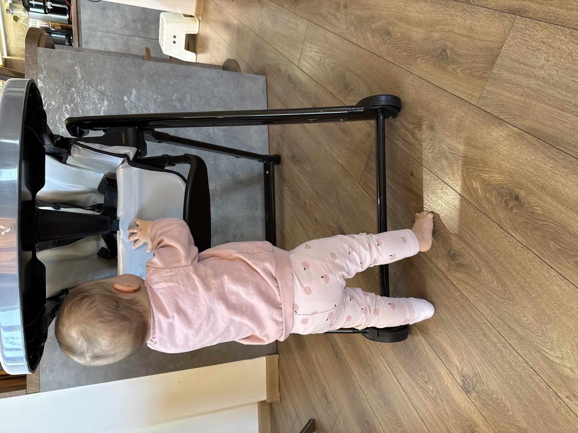 A baby in a pink outfit with a pink long-sleeved shirt and pink pants with a pattern of small white figures is lying on the floor next to a high chair. The high chair has a black frame and a white seat. The floor is made of wood laminate. The location is likely indoors, but the image does not provide enough information to determine the specific location within Sofia, Bulgaria.