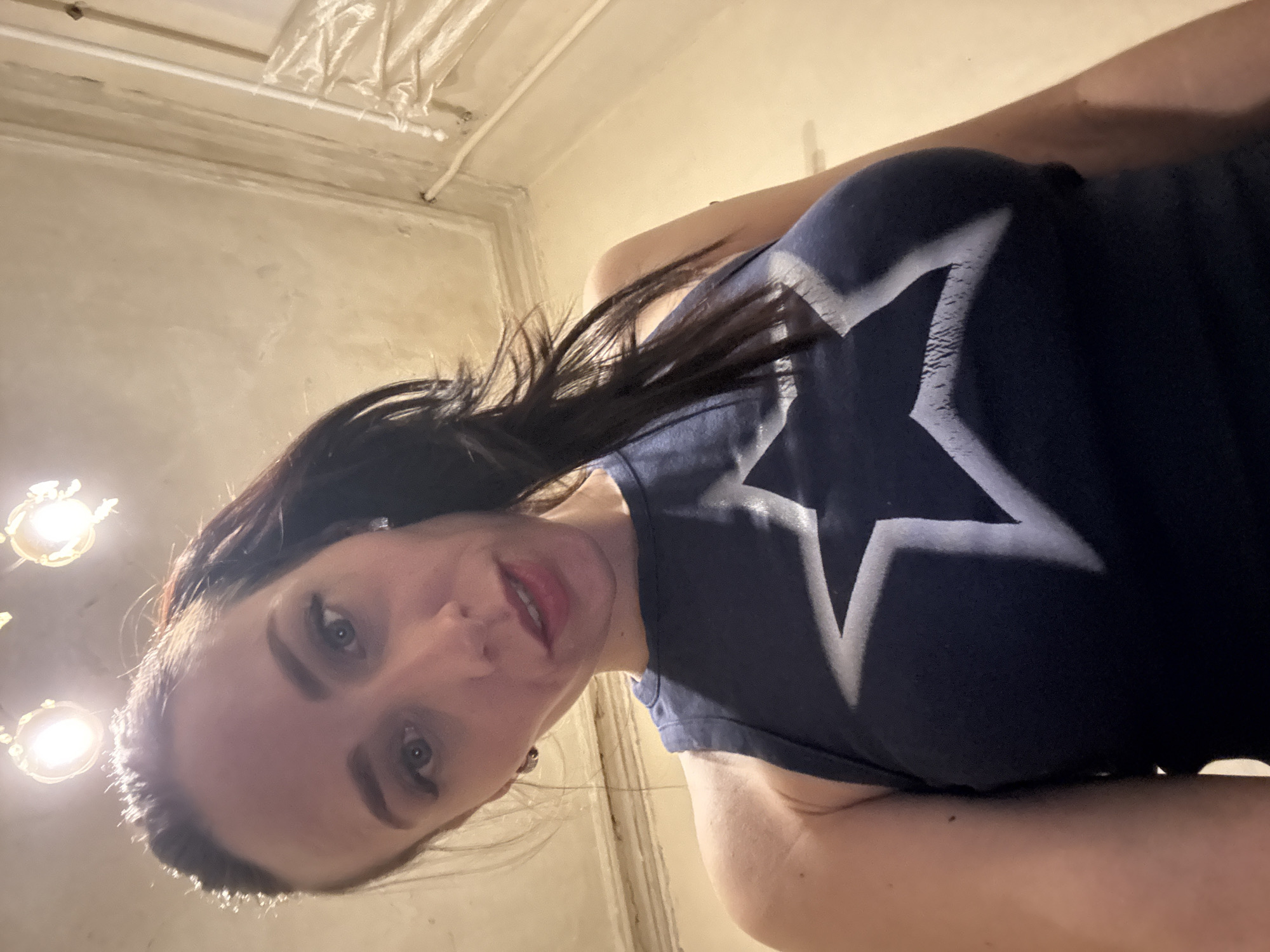 The image captures an upward-angled selfie of a woman with dark hair and blue-gray eyes, looking directly at the camera with a slight, engaging smile. Her head is tilted, and she is wearing red or pink lipstick, with her upper teeth subtly visible. She is dressed in a dark blue sleeveless top adorned with a prominent, faded white outline of a five-pointed star on the chest.
The setting appears to be indoors, against a backdrop of light-colored walls that show textural variations and subtle signs of age. Architectural details, possibly decorative molding or exposed pipes, are visible along the ceiling line. Two light fixtures with bright, exposed bulbs are positioned in the upper left of the frame, providing illumination.
The perspective suggests the woman is looking down at the camera, holding it below her. The indoor lighting indicates no specific time of day or outdoor weather conditions. The general aesthetic of the background, including the style of the walls and ceiling, aligns with the provided location context of Saint Petersburg, Russia, hinting at an older building or residence. There is no visible alphanumeric text, only the star graphic on her clothing.