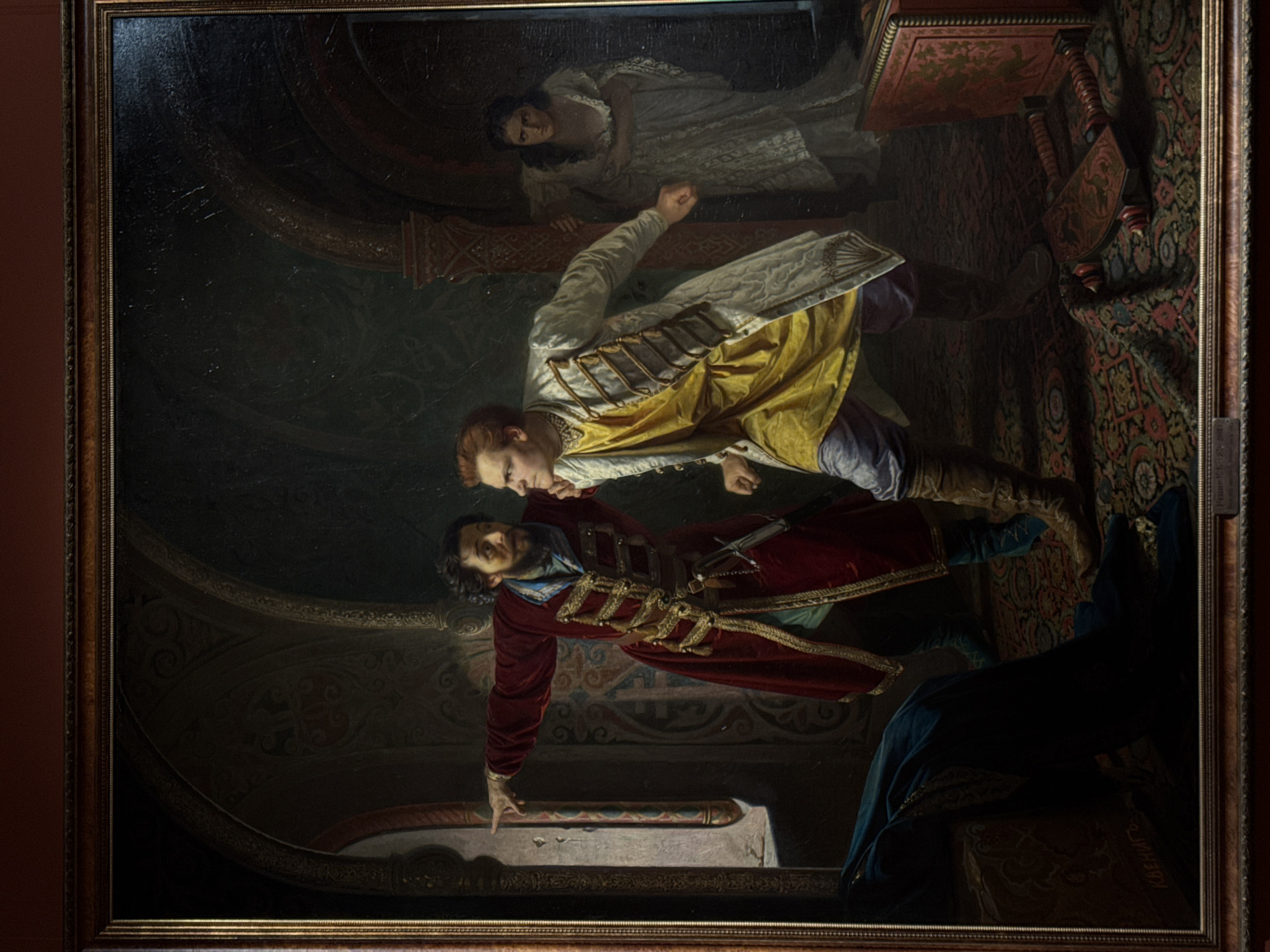 The image shows a painting with a wooden frame, depicting a dramatic scene. A man in a red and gold robe, with a dagger at his side, appears to be grabbing the throat of another man wearing a yellow and white outfit. A woman in a white dress is visible in the background, seemingly observing the event. The scene takes place in an ornate interior with dark walls and patterned carpeting. The painting is titled "Princess Tarakanova". The location is the Nizhny Novgorod State Art Museum in Russia.