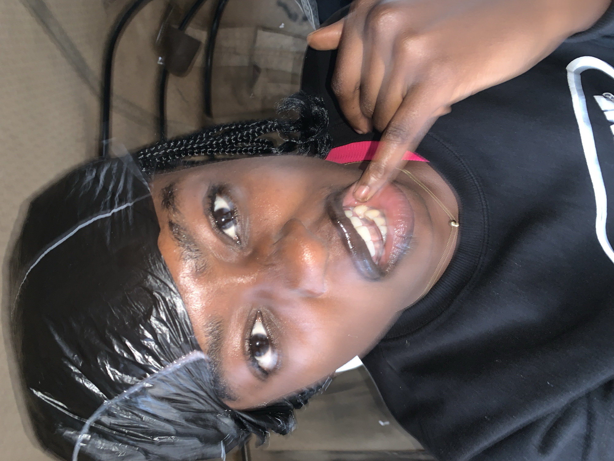 The image shows a close-up of a young Black woman wearing a black durag and a black sweatshirt. She has braids styled down the side of her head, a thin gold necklace, and is wearing a pink strap of some sort visible near her neck. Her mouth is open, and she is touching her teeth with her right index finger. The background is blurry and appears to be indoors, with some dark furniture and what looks like a metallic frame. There is no indication of the city of Jos, Nigeria, within the image or its metadata.